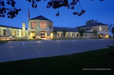 Well-lit modern building at dusk with a central glass entrance, stone facade, and empty parking lot
