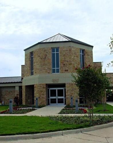 Brick building entrance with glass doors, a central tower, and landscaped lawn under a cloudy sky