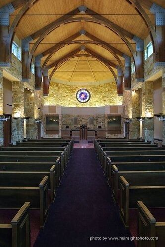 Empty church sanctuary with wooden vaulted ceiling, pews, purple carpet, and circular stained-glass window.