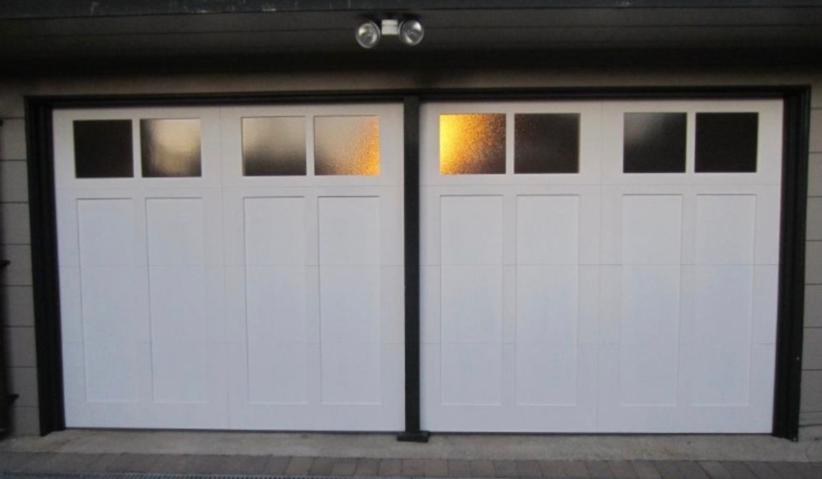 Two white garage doors with small windows, lit from inside at dusk