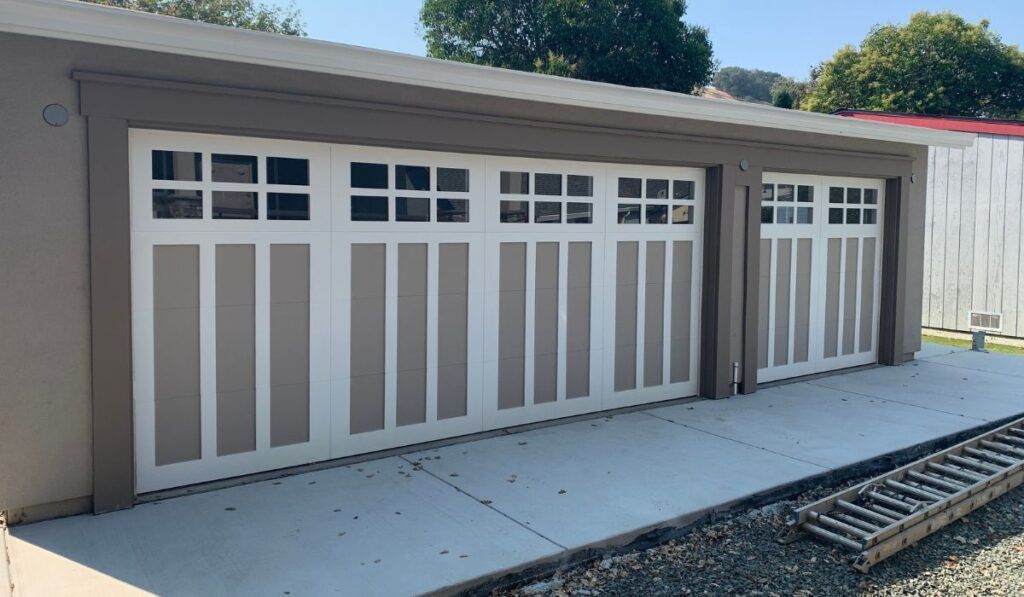 White garage doors on a beige building with small windows, beside a concrete driveway and gravel border.
