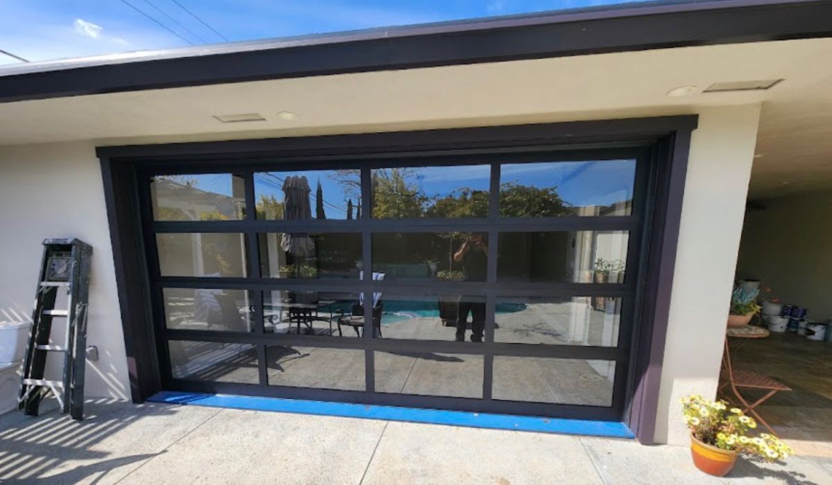 Black-framed glass garage door on a white house, with blue painter’s tape at the base and a ladder nearby.