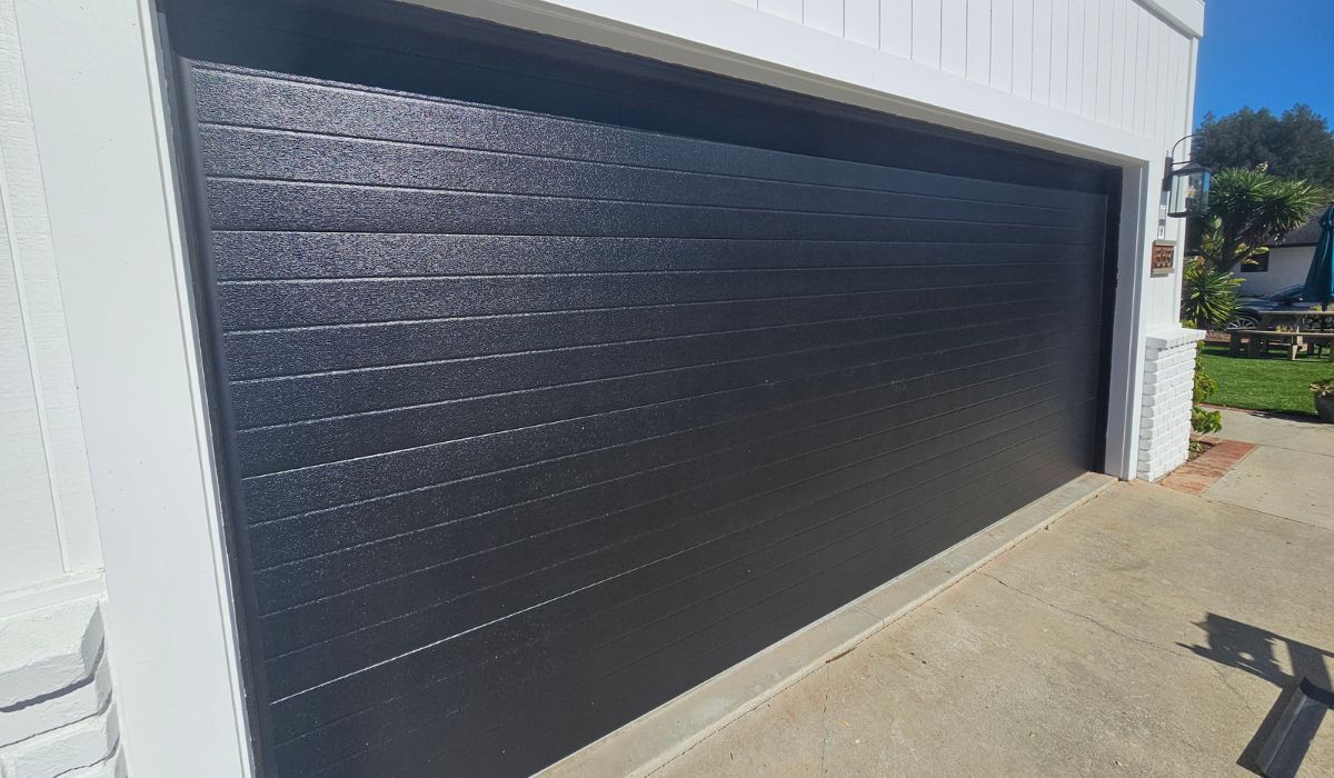 Black garage door on a white building beside a concrete driveway in bright sunlight