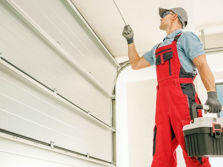 Technician in red overalls inspecting a garage door track with a drill and toolbox