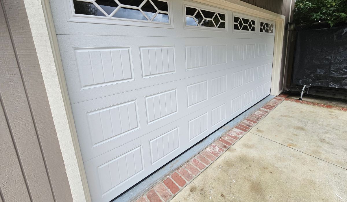 White garage door with decorative windows beside a concrete driveway and brick border