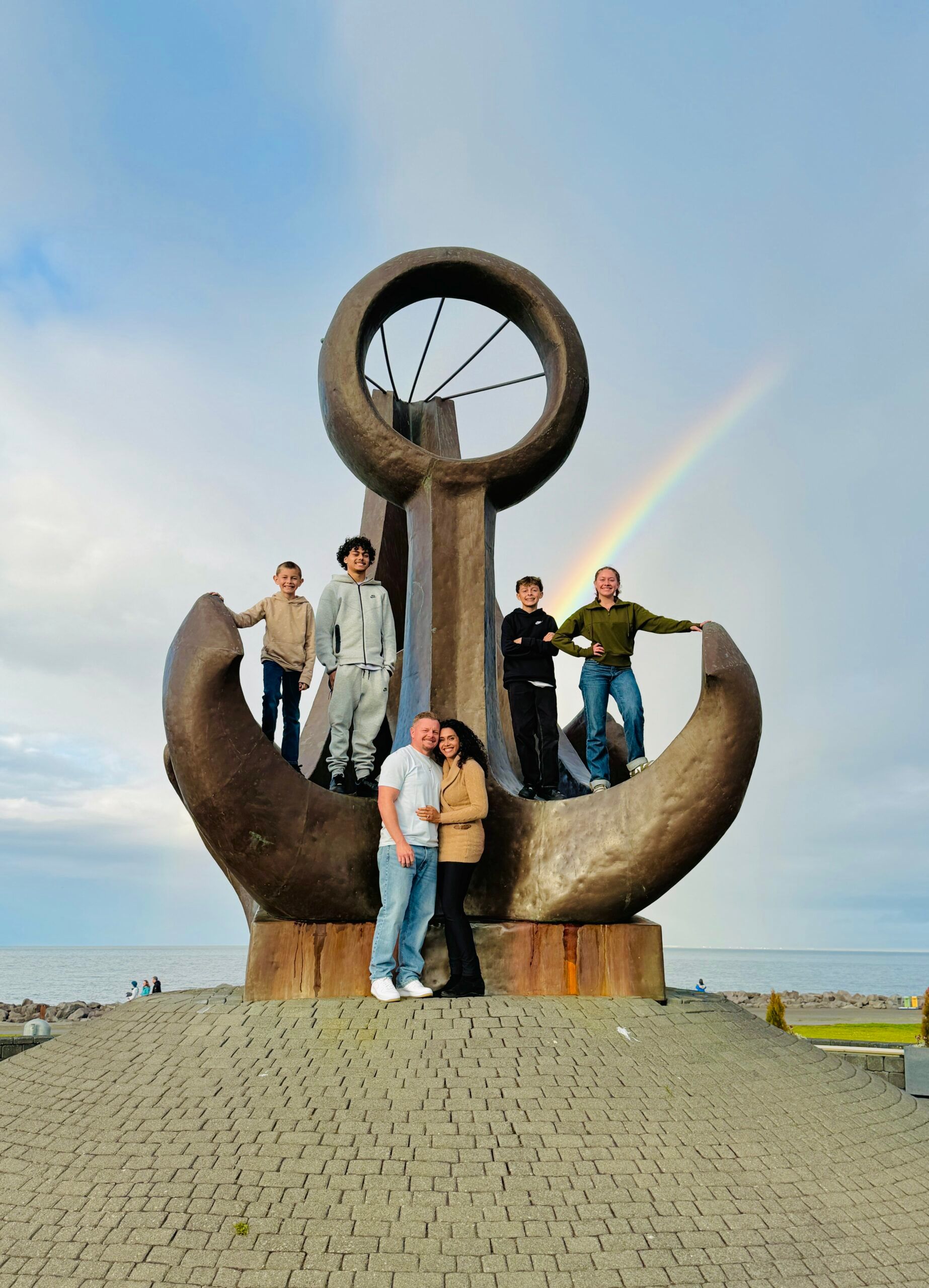 Group posing on a large metal anchor sculpture outdoors under a cloudy sky.