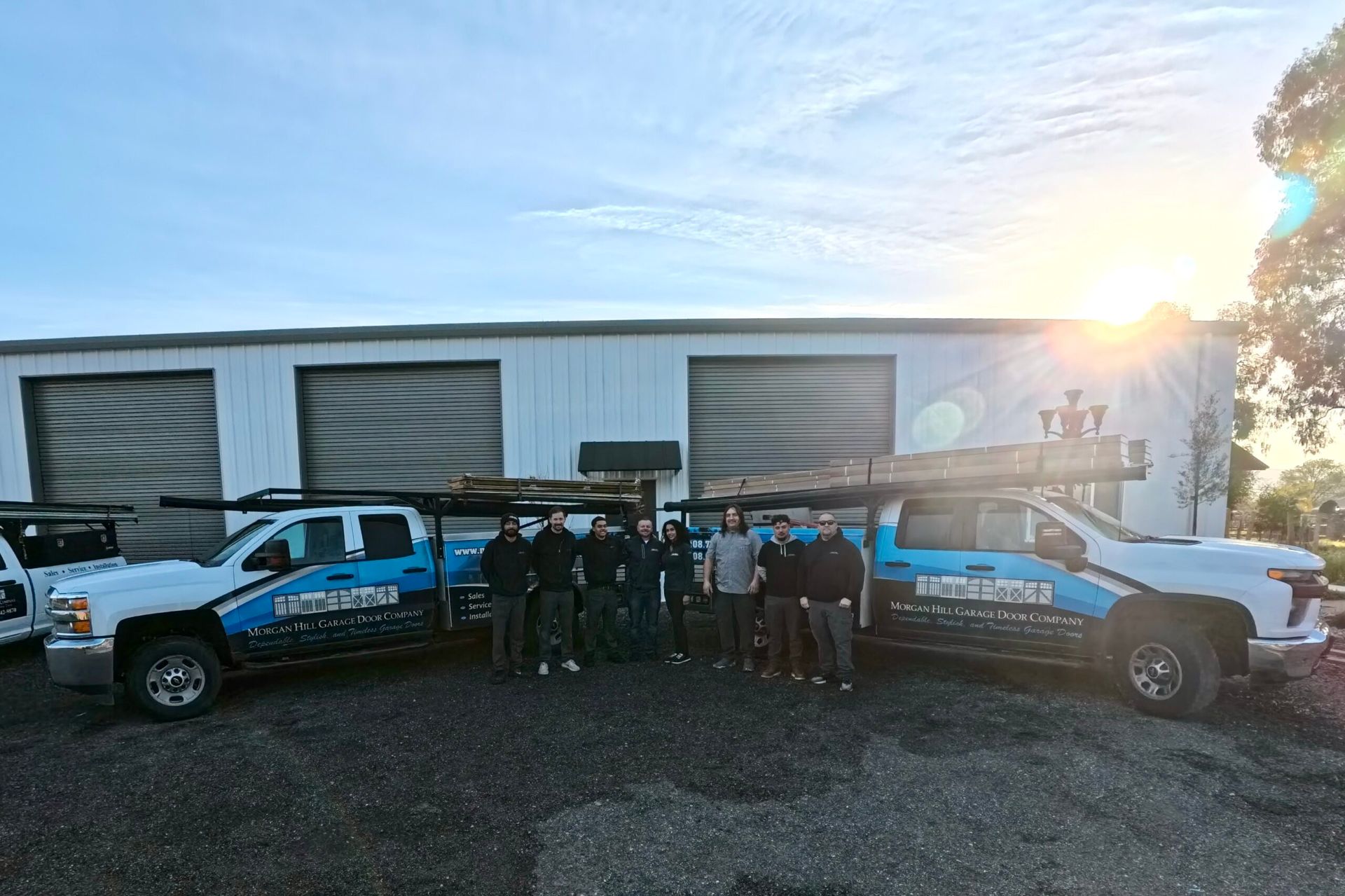 Two blue utility trucks parked outside a warehouse with a group standing between them at sunset