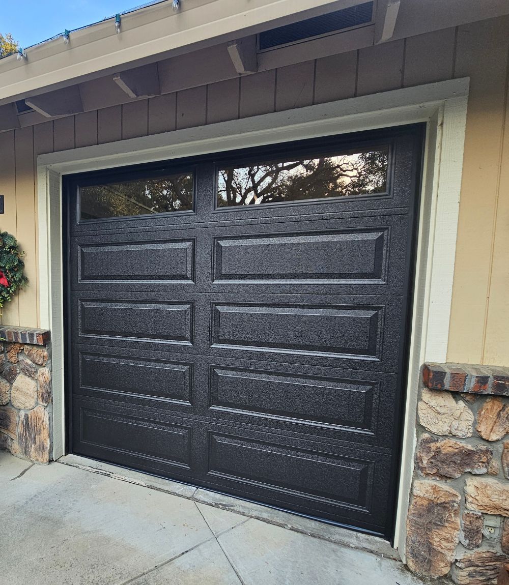 Black textured double garage door framed by tan siding and stone accents under a roof overhang
