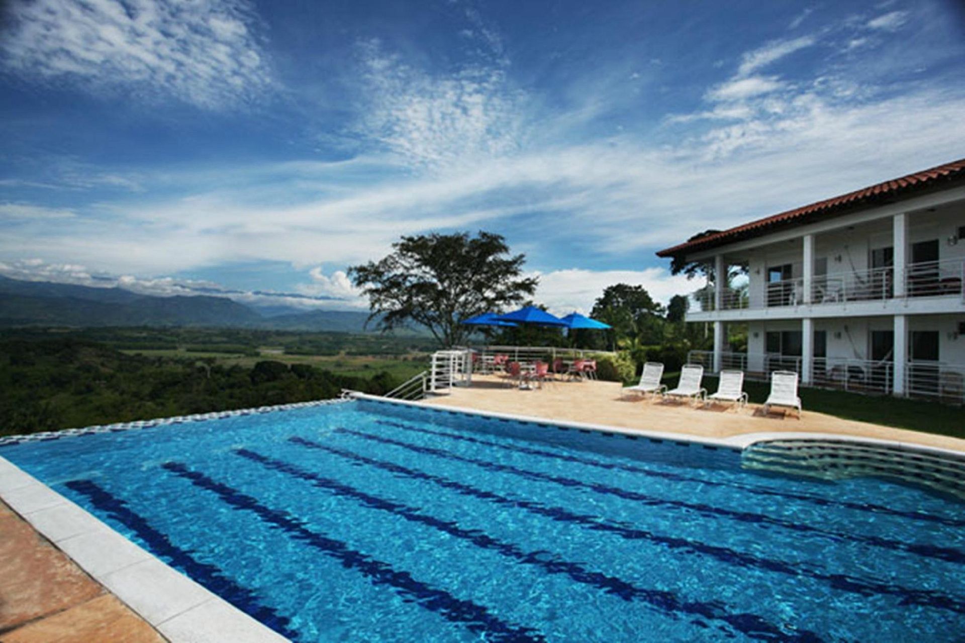 Una gran piscina está rodeada de sillas y sombrillas frente a una casa.