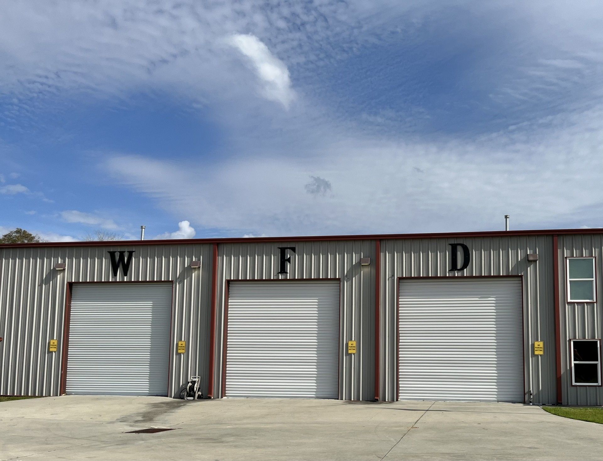 Industrial building with three white garage doors labeled W, F, and D under a cloudy blue sky
