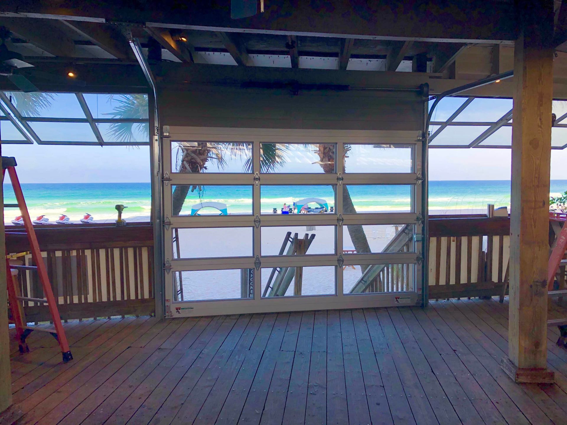 Beachfront patio with wooden deck and railings, framing a bright turquoise ocean and sandy shore beyond.