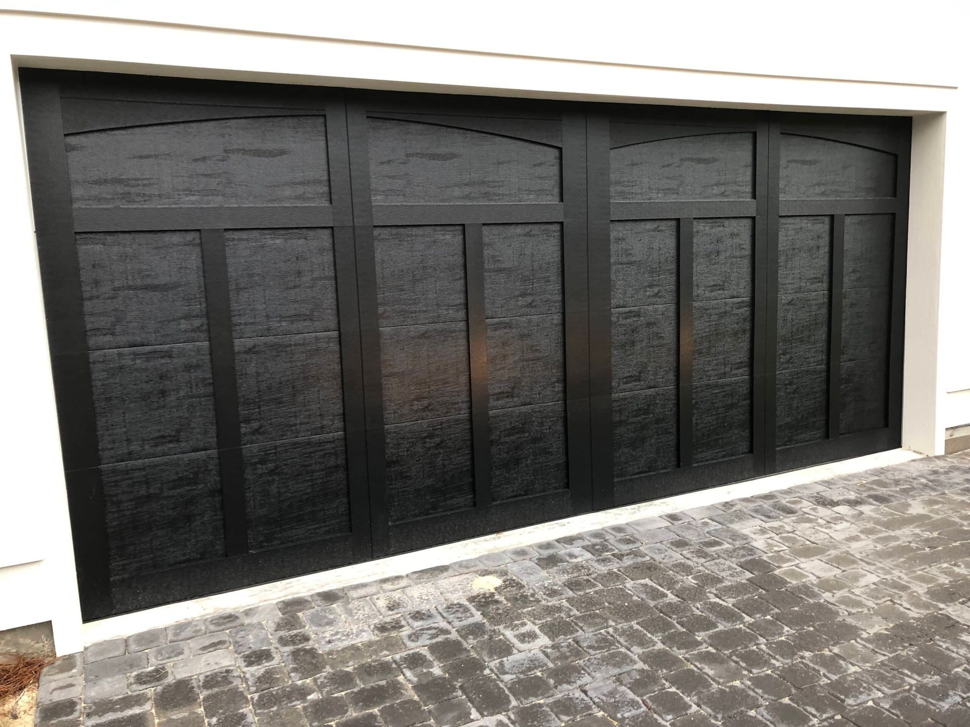 Black modern garage door with vertical panels set in a white wall above a cobblestone driveway