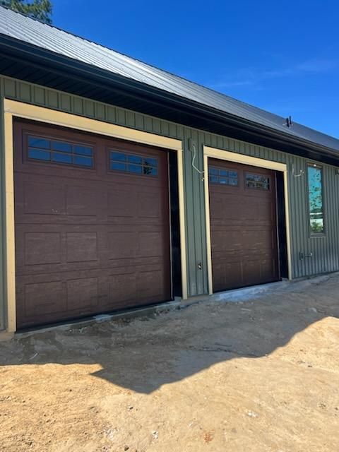 Two brown garage doors on a dark-sided house with tan trim and sandy ground in front