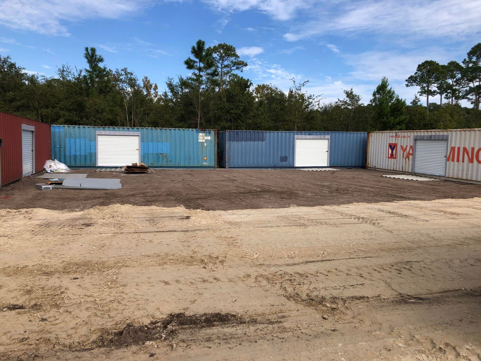 Row of shipping containers in a dirt lot beside trees under a blue sky