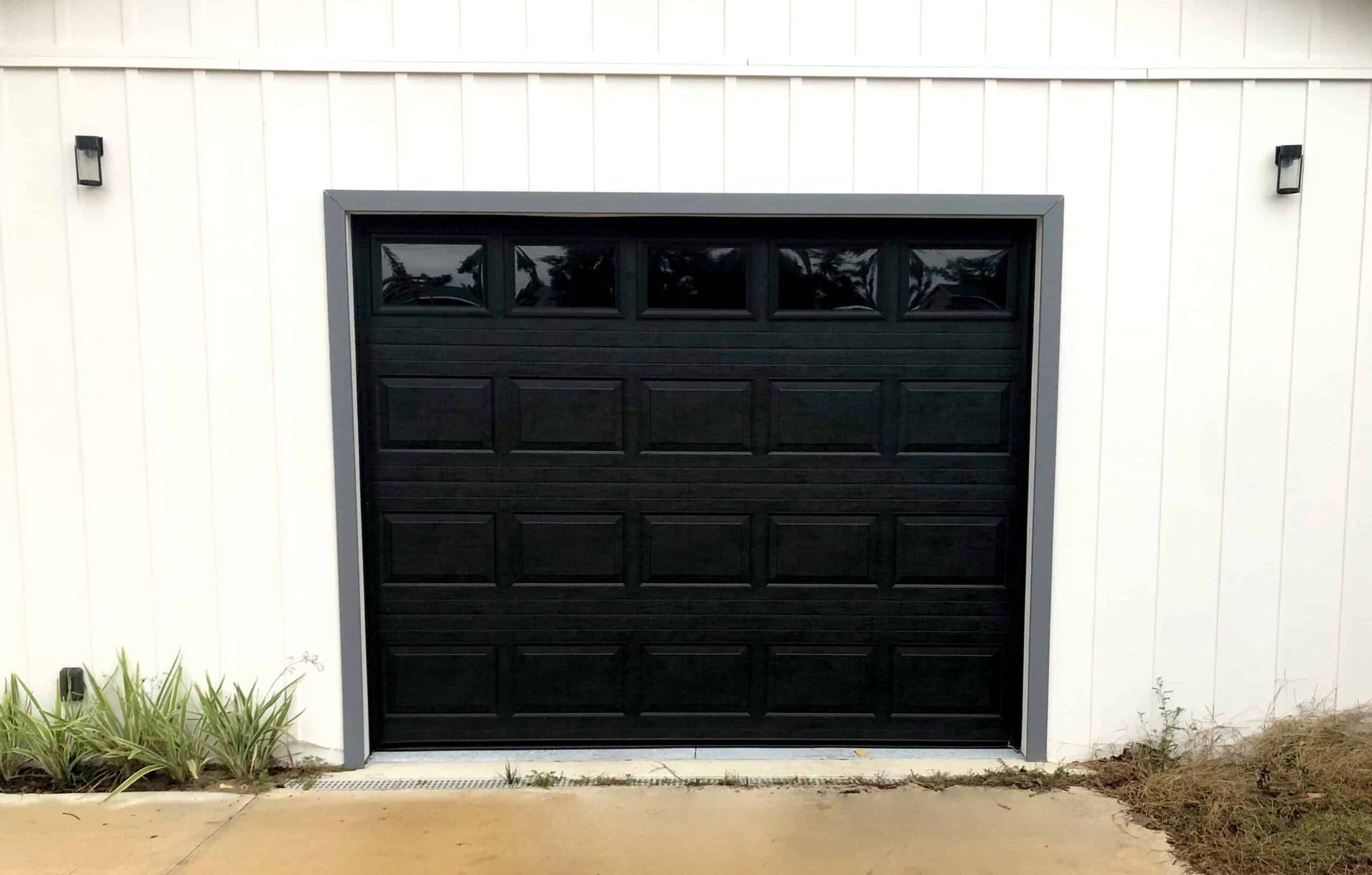 Black garage door centered on a white exterior wall with small windows and plants on the left.