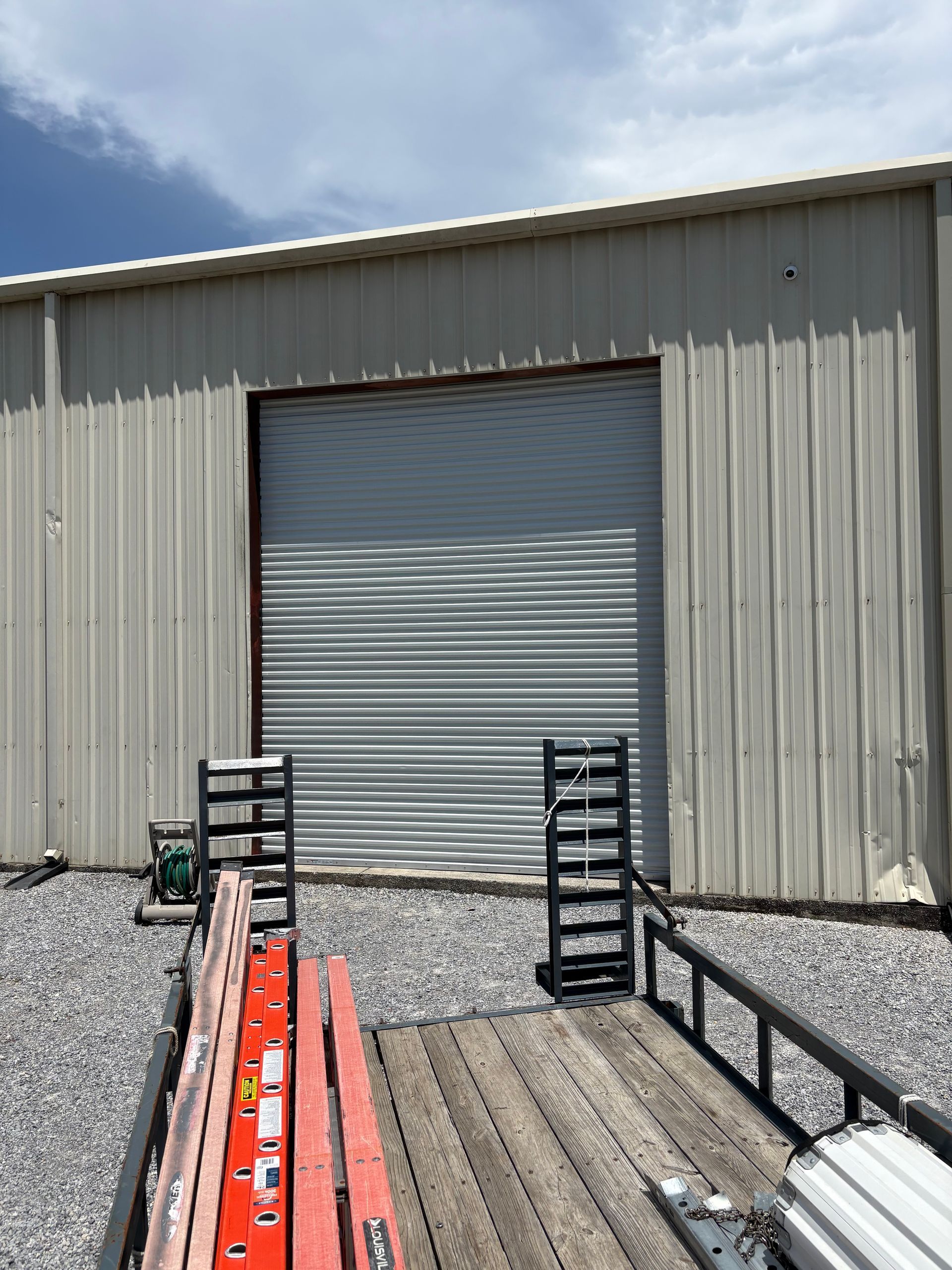 Industrial building with a closed gray roll-up door and metal ramps over gravel under a partly cloudy sky