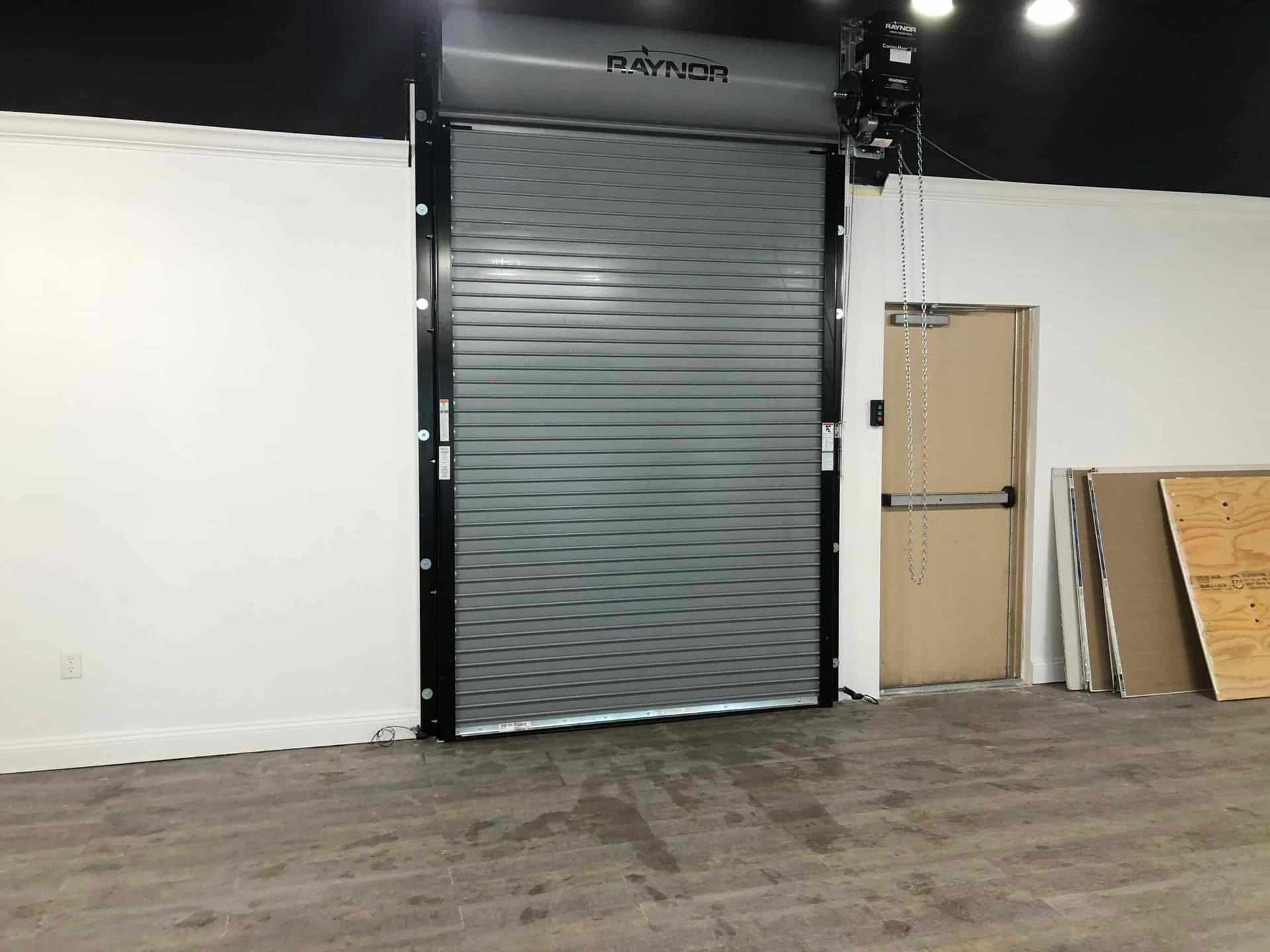 Empty warehouse with a closed gray roll-up door, white wall, and stacked boards on the right.