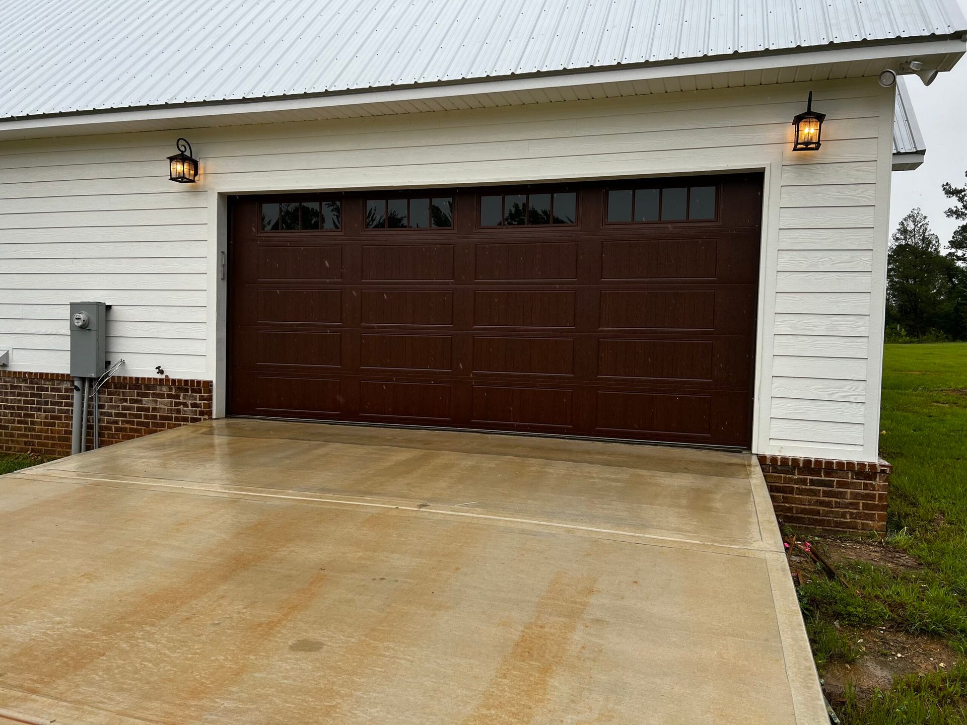 Brown double garage doors on a white garage with lit wall lanterns and a concrete driveway