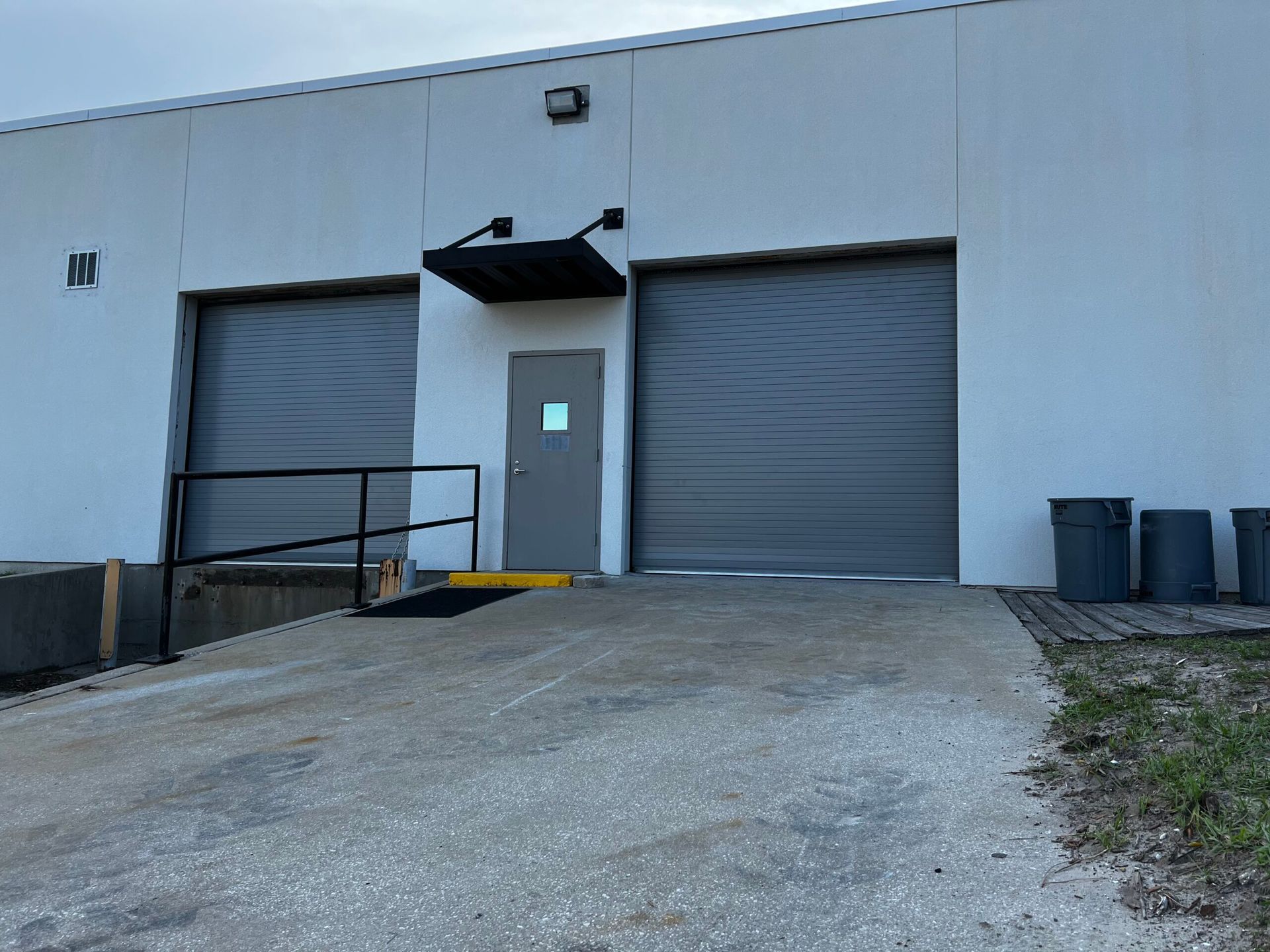 Industrial warehouse exterior with two roll-up doors, a central entrance, and a gravel loading ramp.