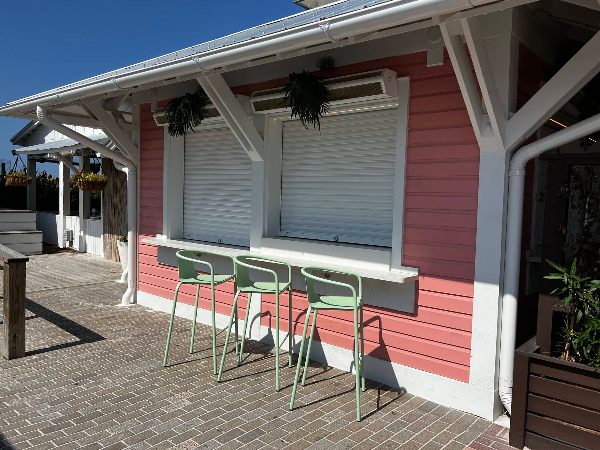 Pink storefront with white shutters, hanging plants, and green bar stools on a sunlit patio