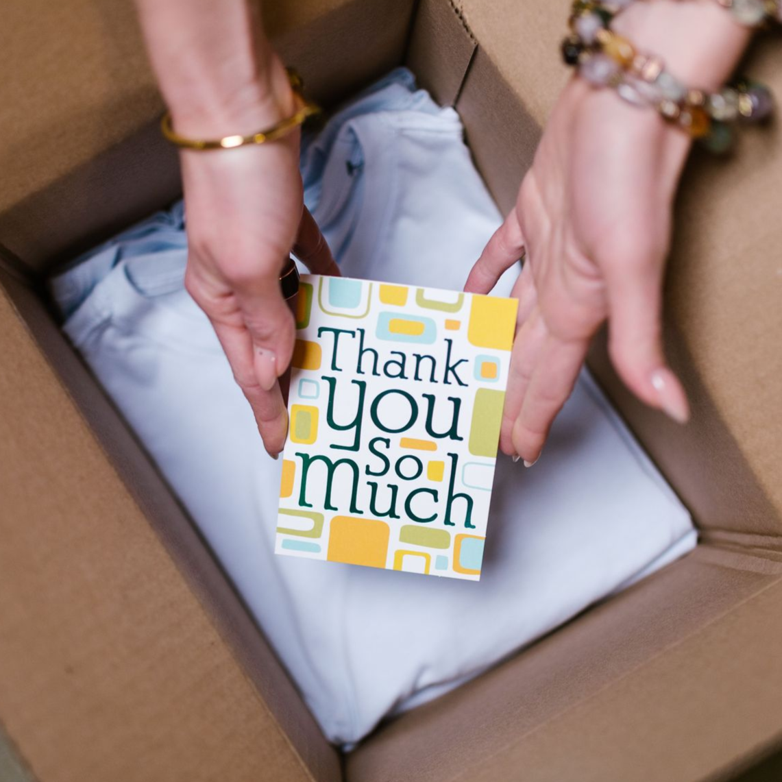 Hands placing a “Thank you so much” card on a wrapped gift in a box