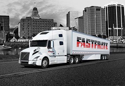 White Fastenal semi-truck driving on a road with a city skyline in the background