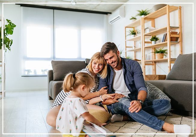Family of three, sitting on floor, looking at book. Smiling, happy expressions. Living room setting.