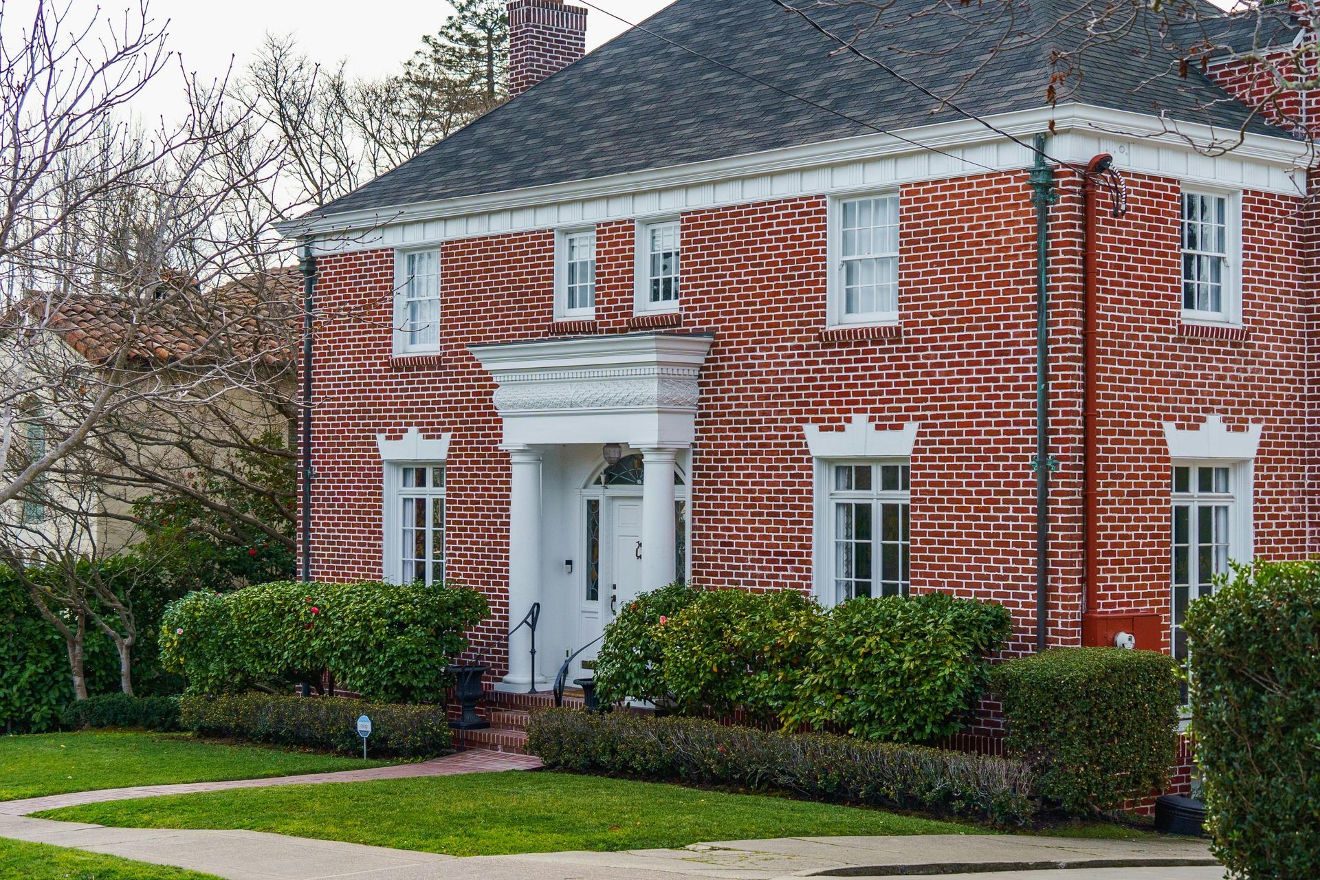 Red brick two-story house with white trim, front porch, and manicured hedges in a suburban neighborhood