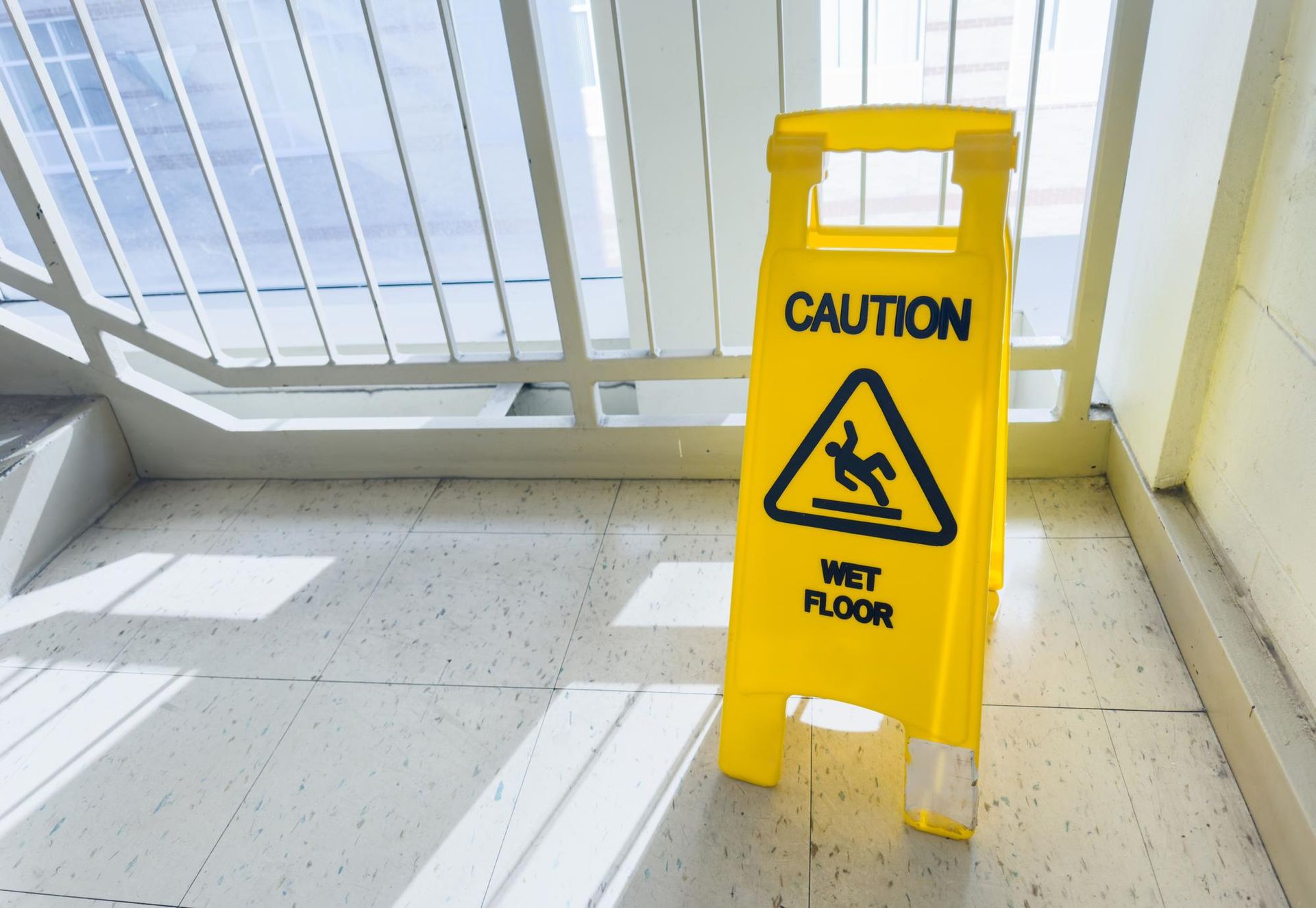 Yellow caution sign on a wet floor near a bright windowed corridor.