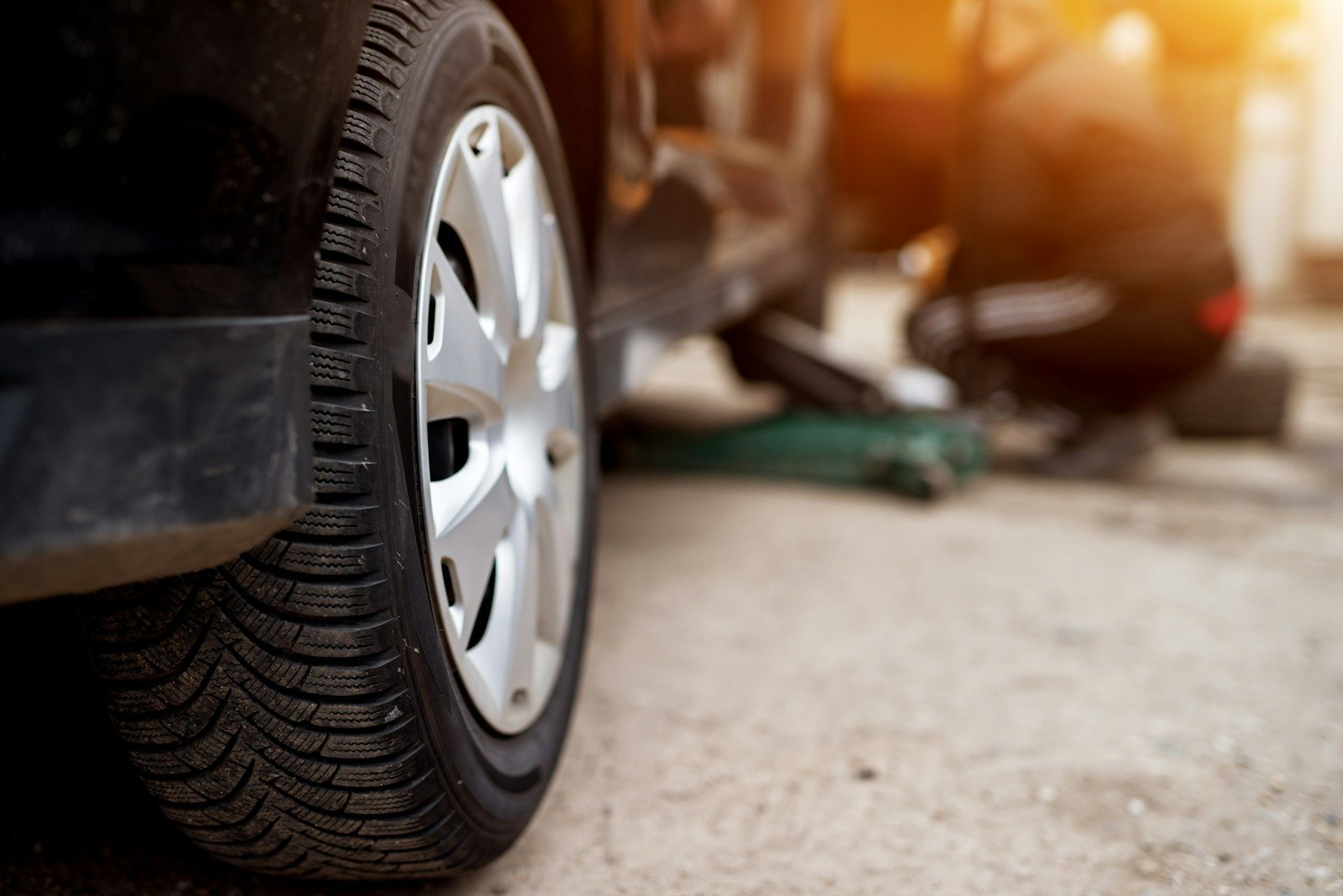 Flat tire on a black car with a person kneeling beside it on a roadside at sunset