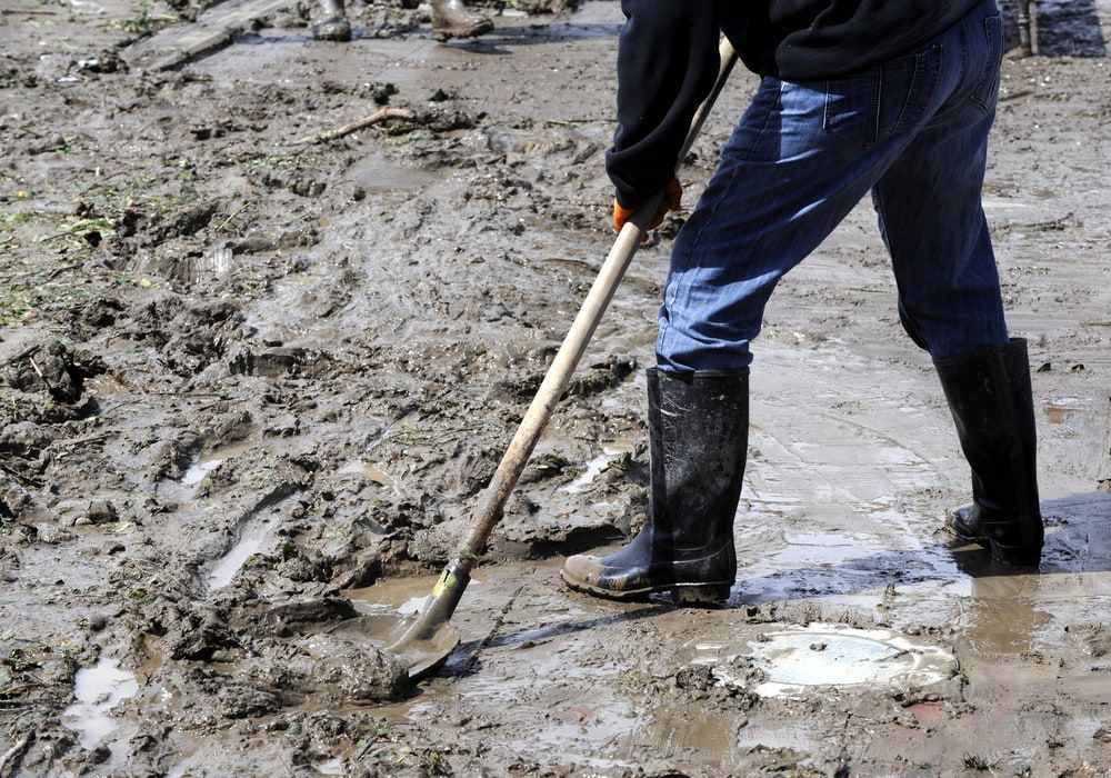 Person in Boots Shoveling Mud From a Muddy Surface Outdoors — Cleanaroo Property Services in Conder, ACT