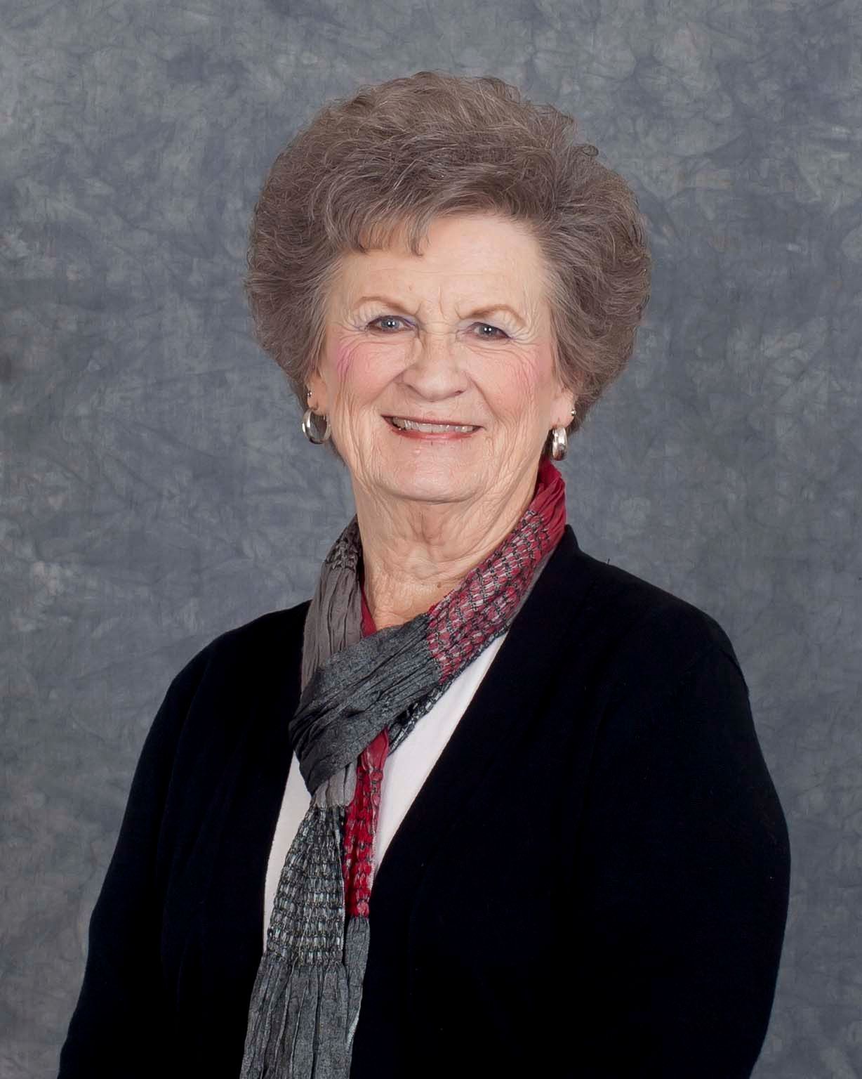 Woman with curly brown hair, smiling, wearing a black sweater and patterned scarf, standing in front of a neutral backdrop.