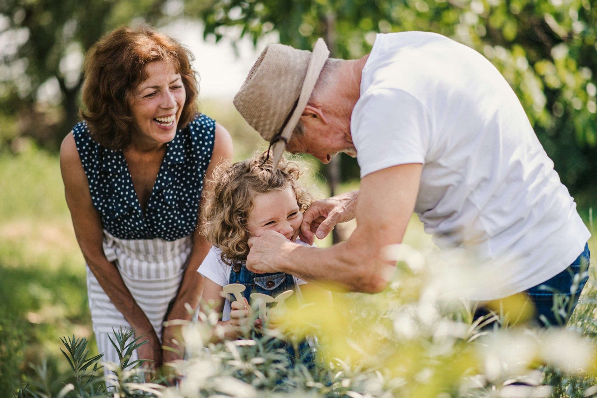 Grandparents playfully interact with a child in a garden; the woman laughs, the man wears a hat.