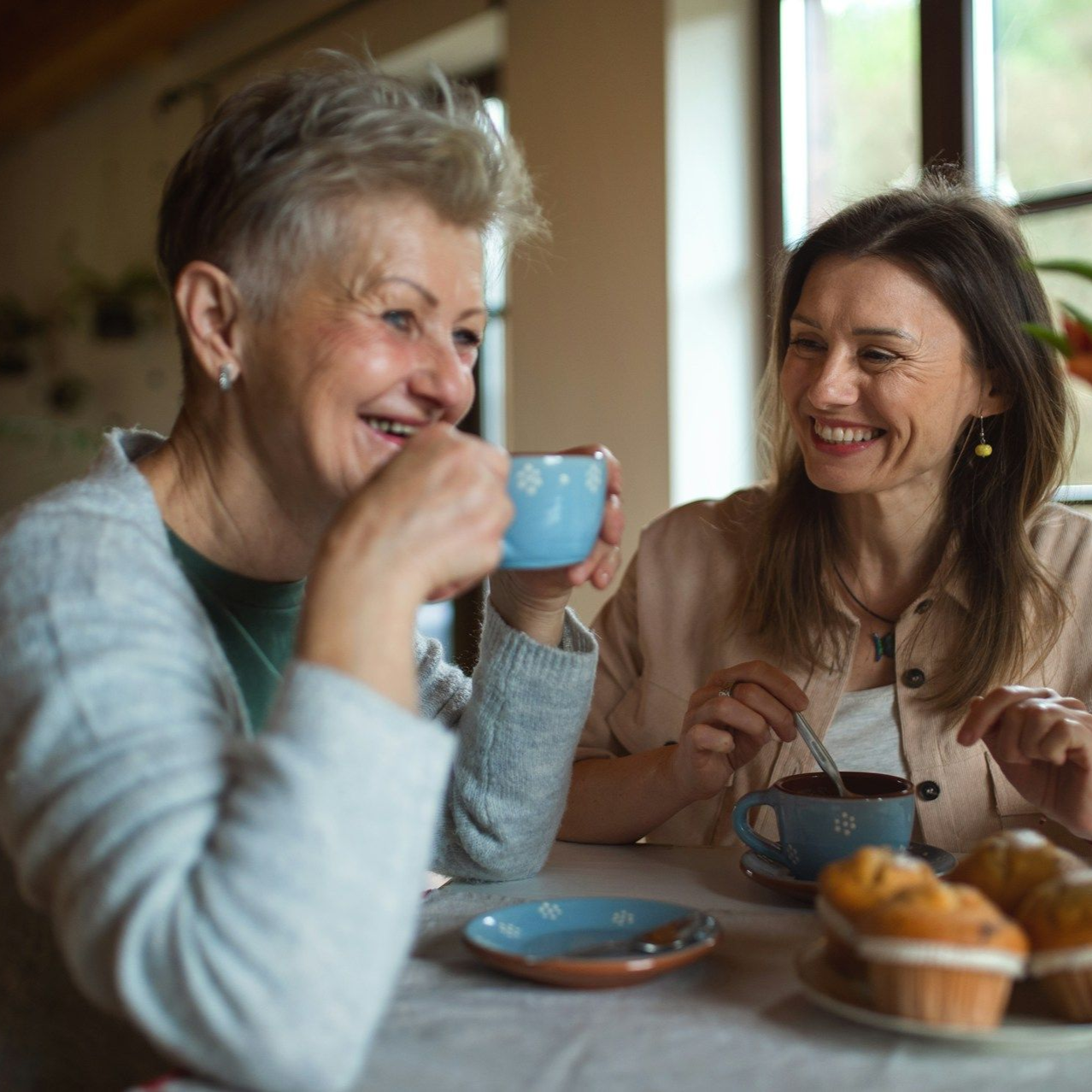 Two women laughing over coffee, muffins on a table. One holds a blue cup, other stirs hers.