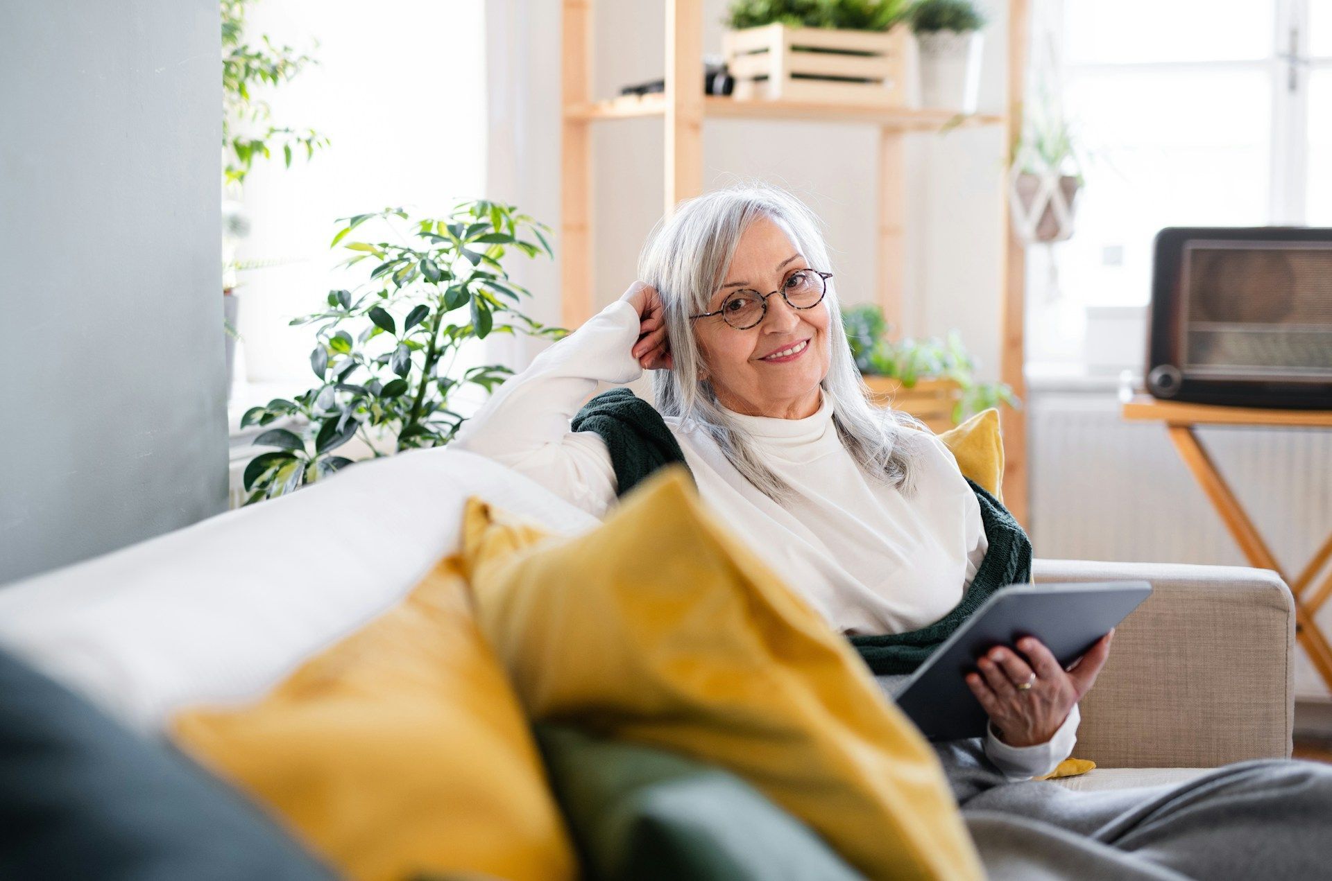 Woman on a couch holding a tablet, smiling. Natural light, plants, and a vintage radio in the background.