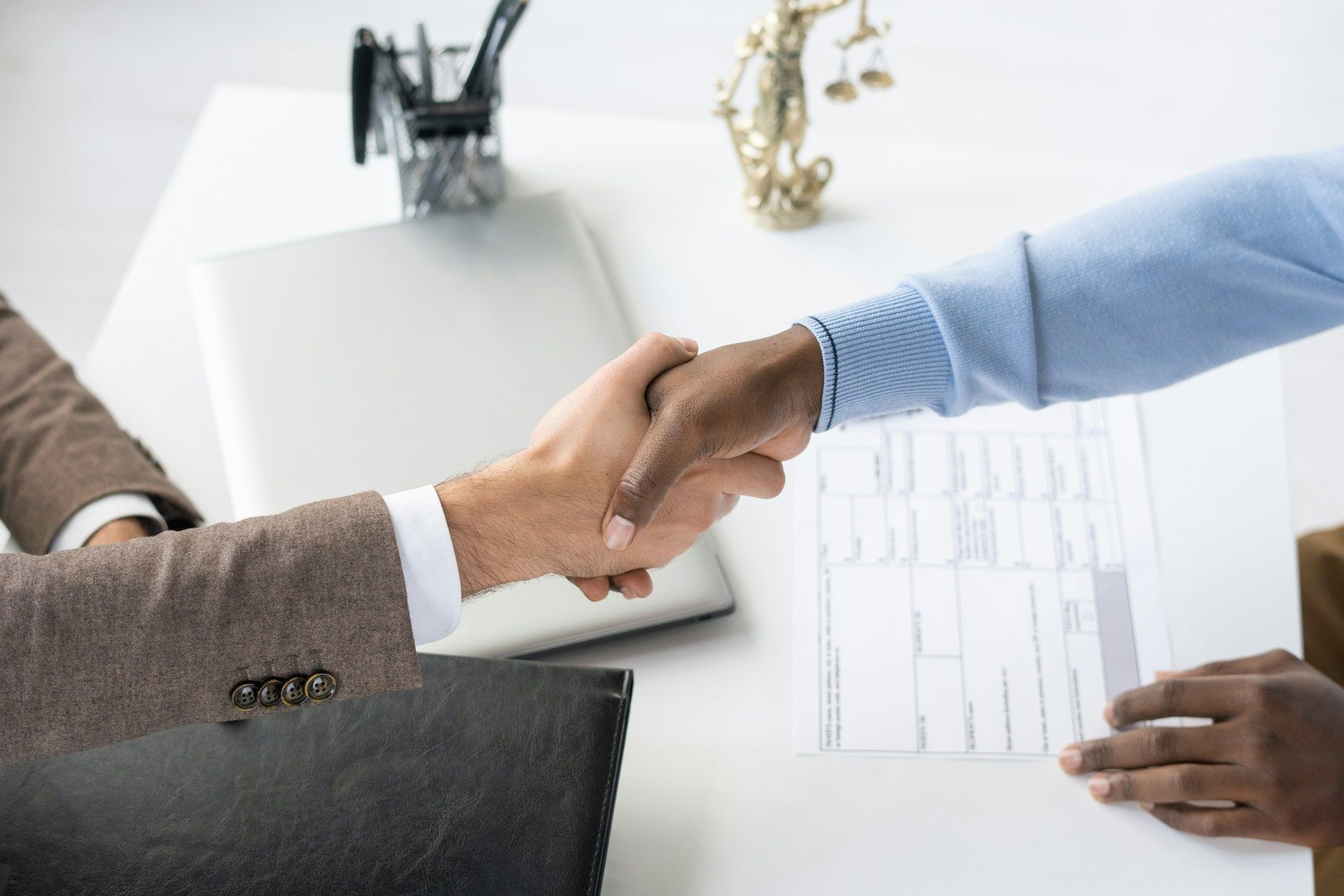 Two people shaking hands over a table with documents and a laptop, possibly a business deal.