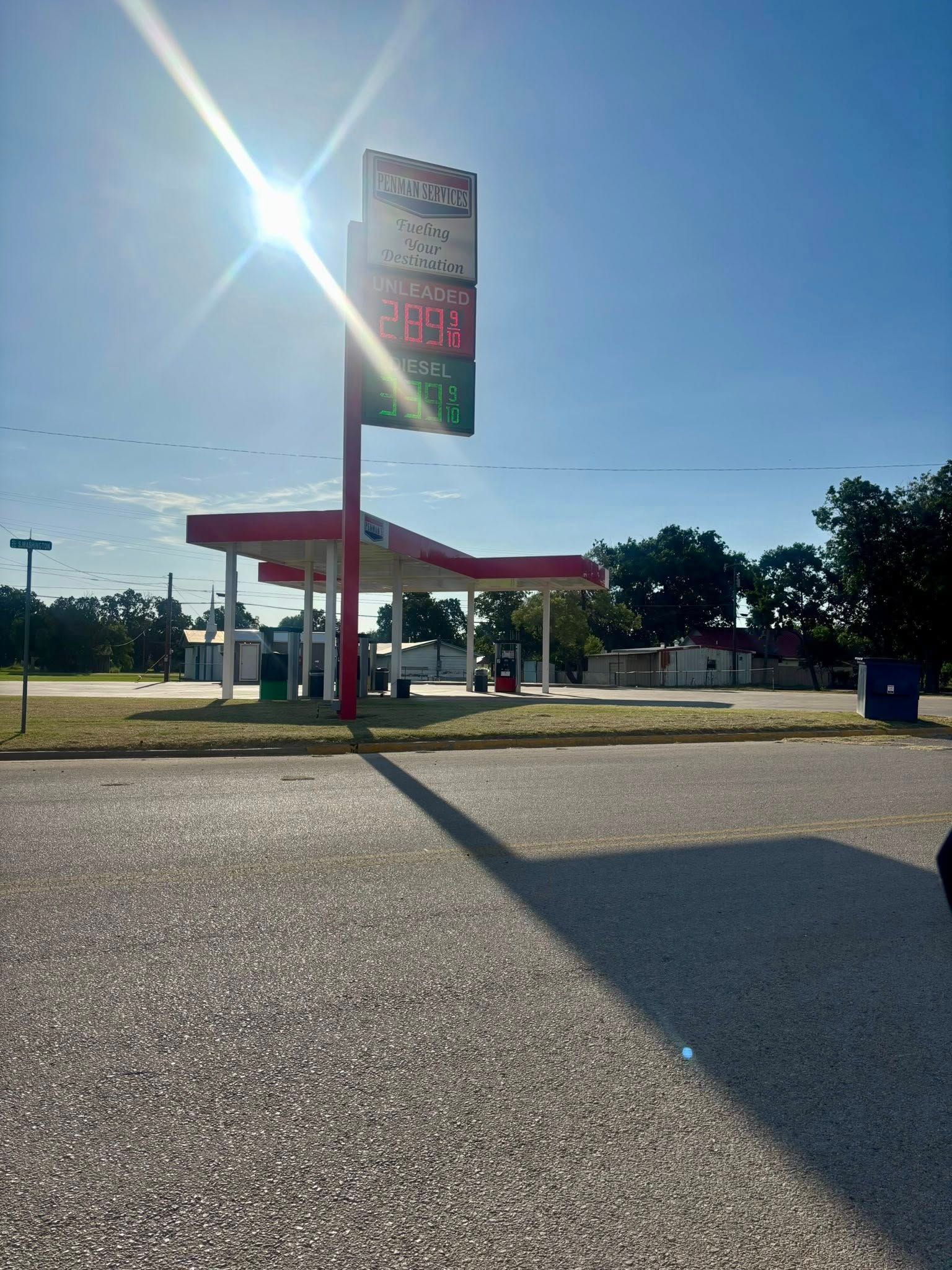 Gas station under a bright sun; red and white canopy, fuel price sign. Blue sky and gravel foreground.