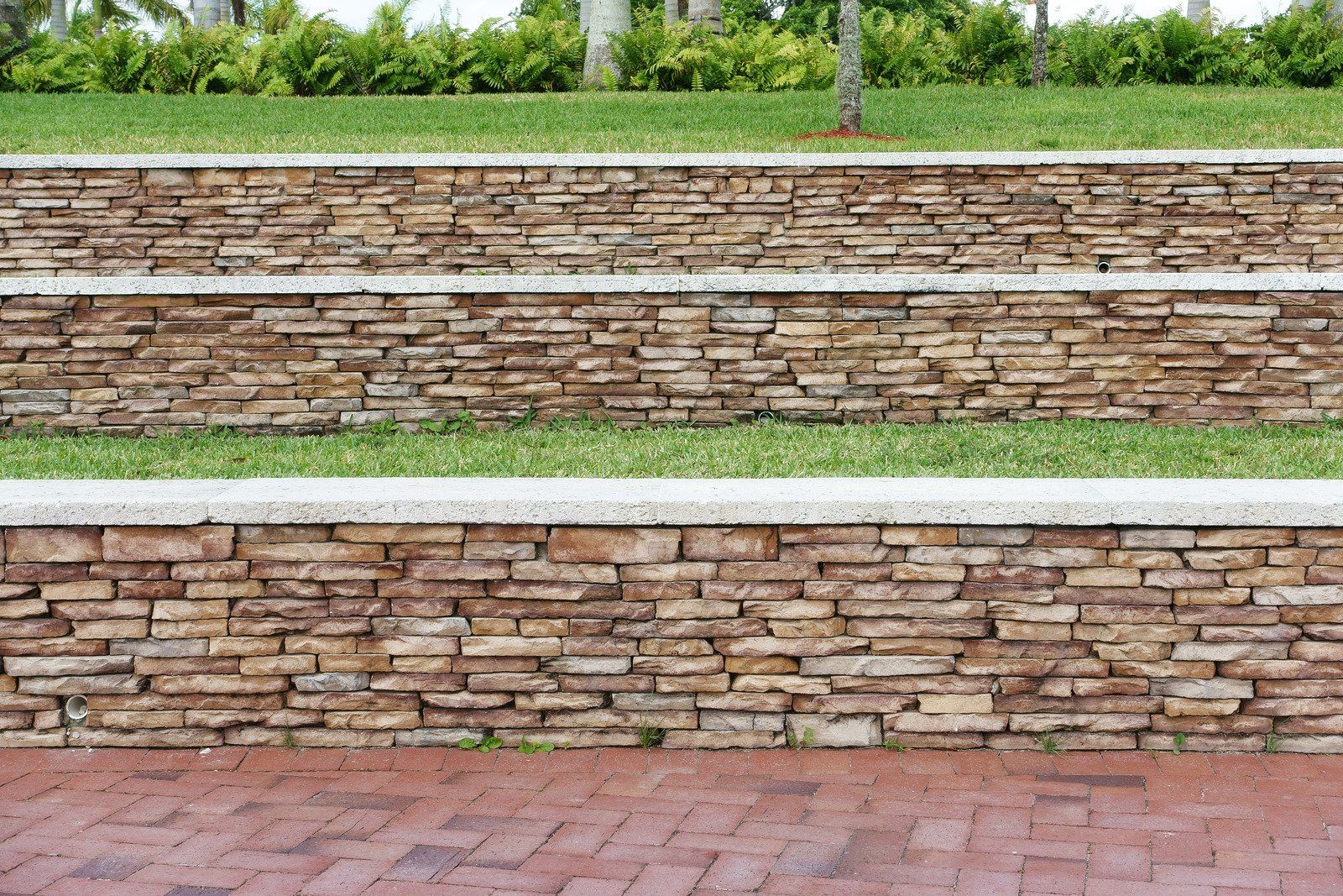 Stone tiered retaining walls with green grass and shrubs on top. Brick walkway in foreground.