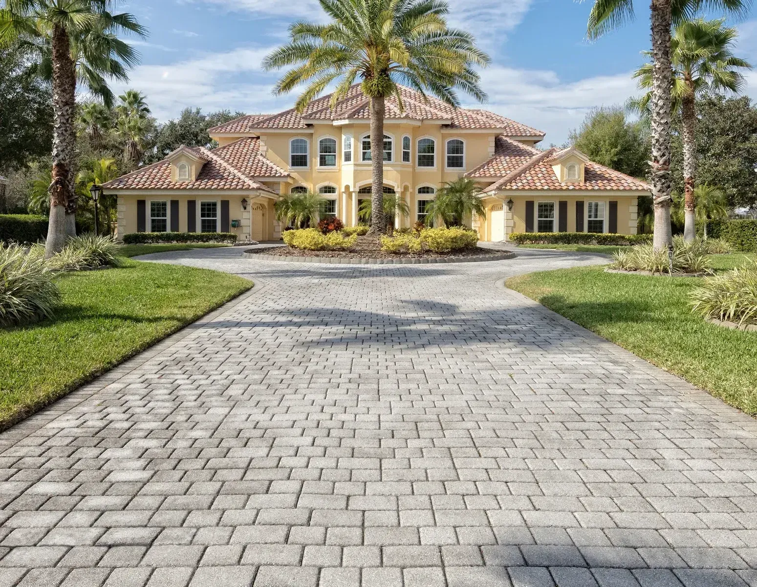 Brick driveway leads to a yellow mansion with red tile roof, palm trees, and green lawn.