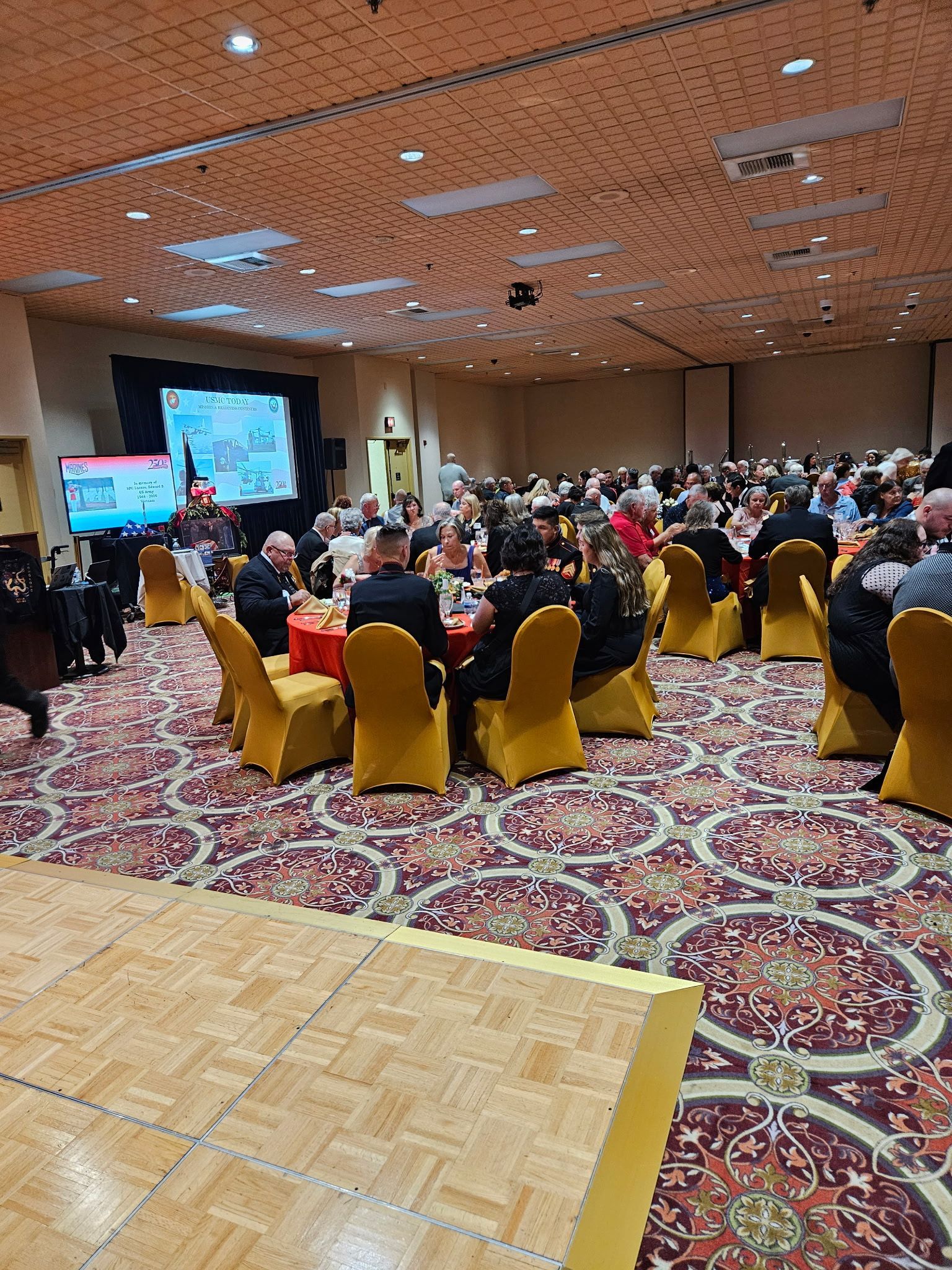 Conference audience seated at round tables in a patterned hall, facing a presentation screen on stage.