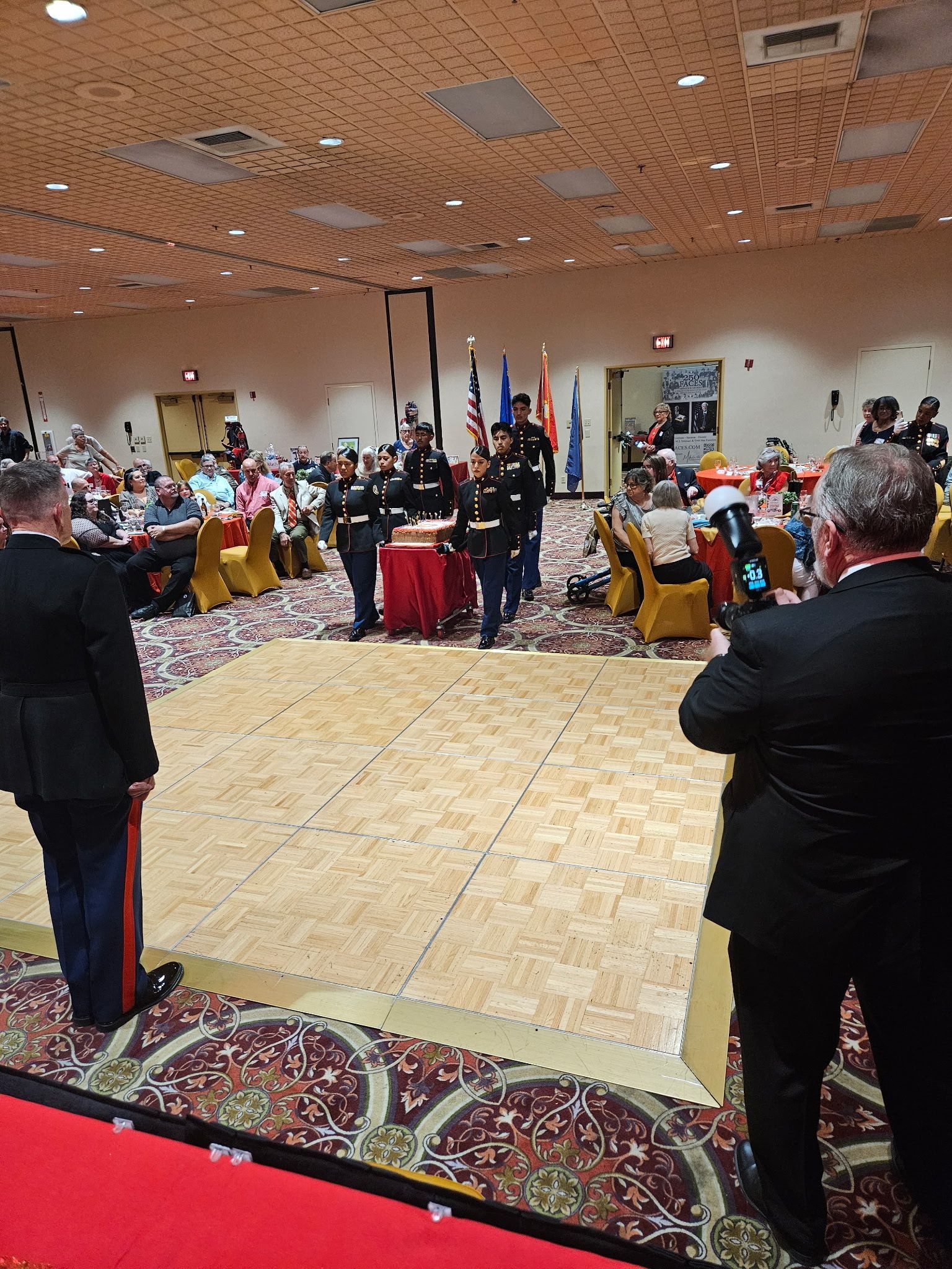 People seated around a banquet hall dance floor during a formal indoor event.