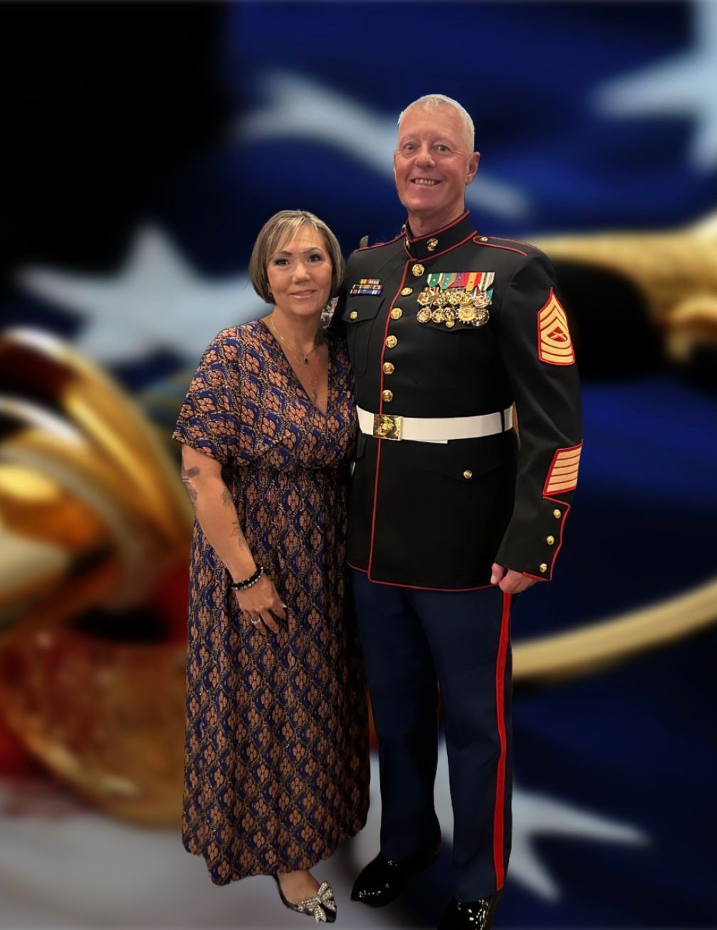 Woman posing beside a man in a dress military uniform, both smiling against a blurred flag backdrop.