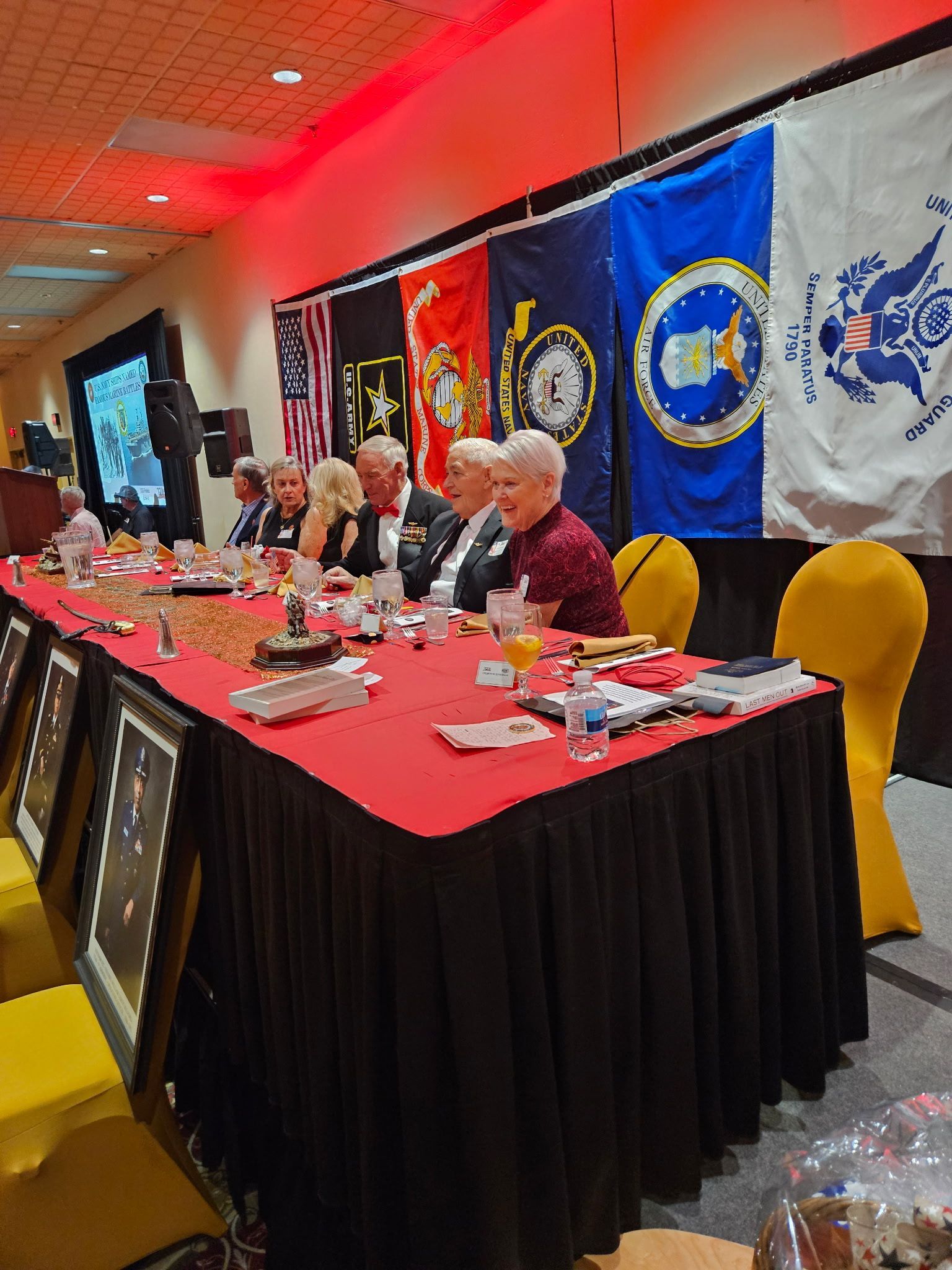 Panelists seated at a red-draped table under military service banners in a conference room