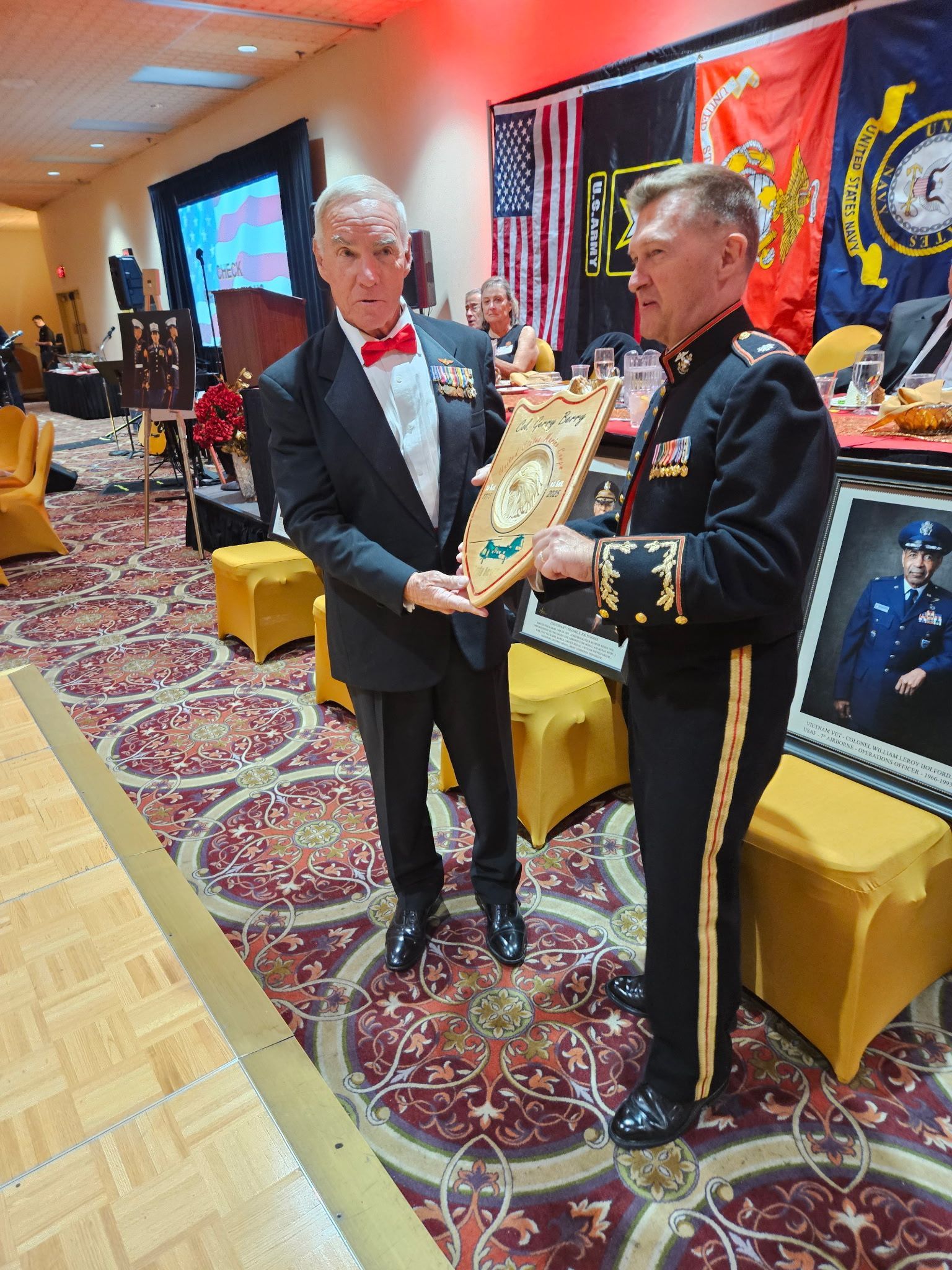 Two men in formal attire exchange a framed plaque at a decorated event with flags and display tables.