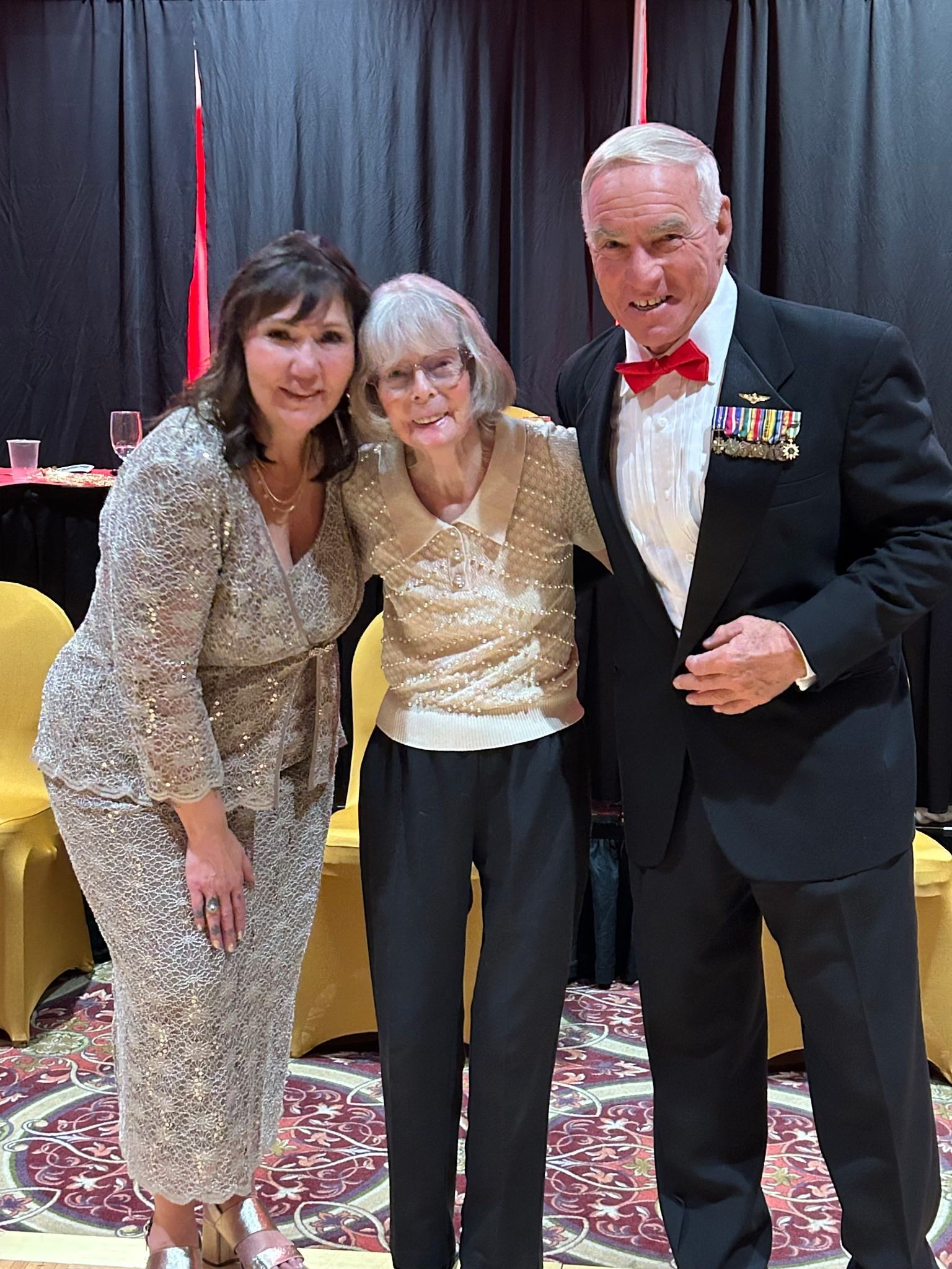 Three people posing together at a formal event, with black drapes and a red carpet in the background