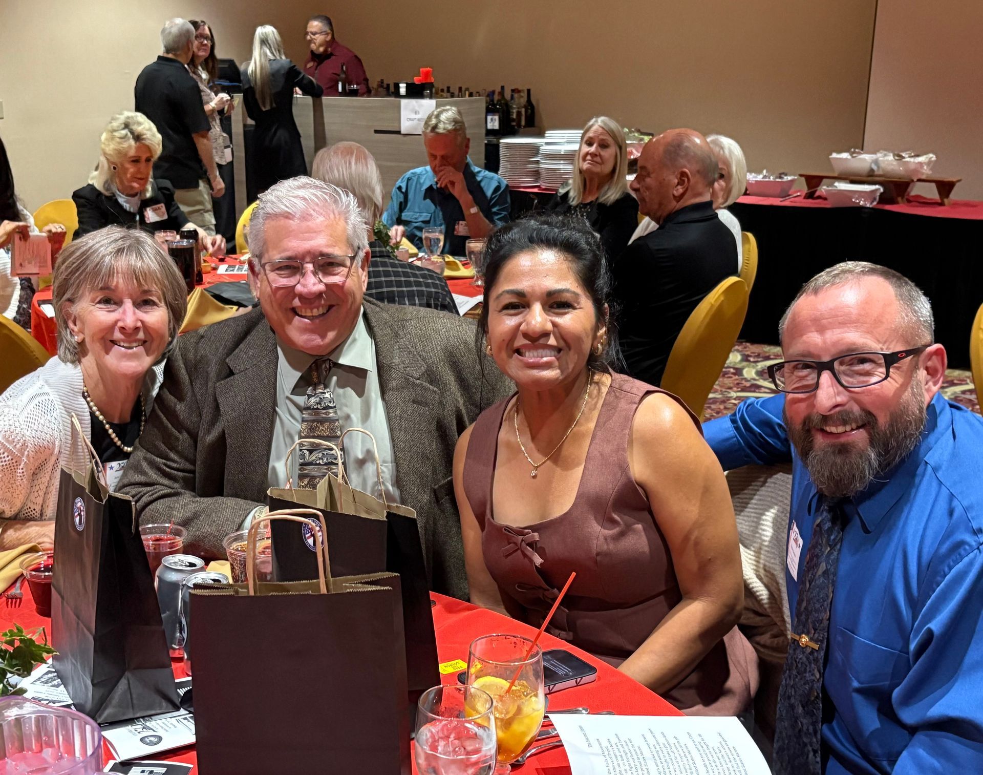 Four people smile at a restaurant table with drinks and a handbag in the foreground.