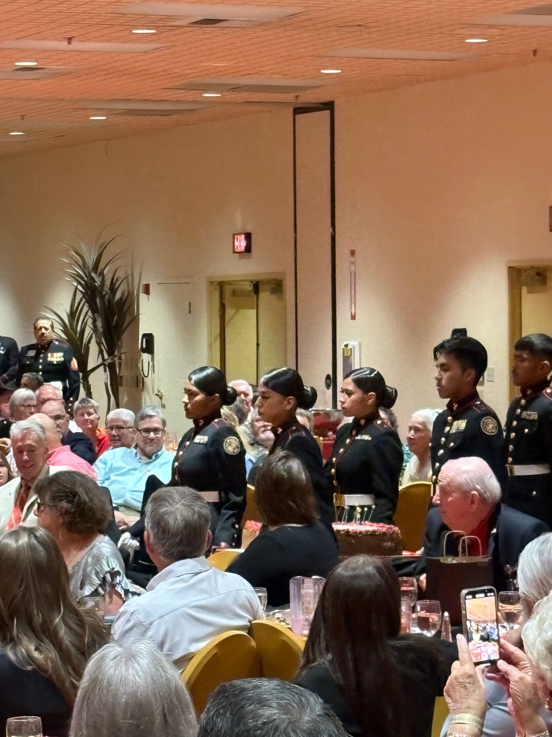 Uniformed officers stand in a banquet hall aisle as seated guests look on.