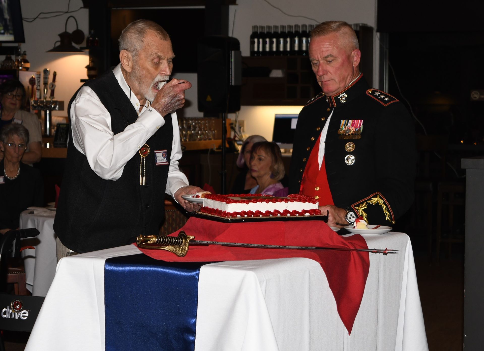 Two men in military uniforms cut a red-white-blue layered cake at a table during a ceremony.