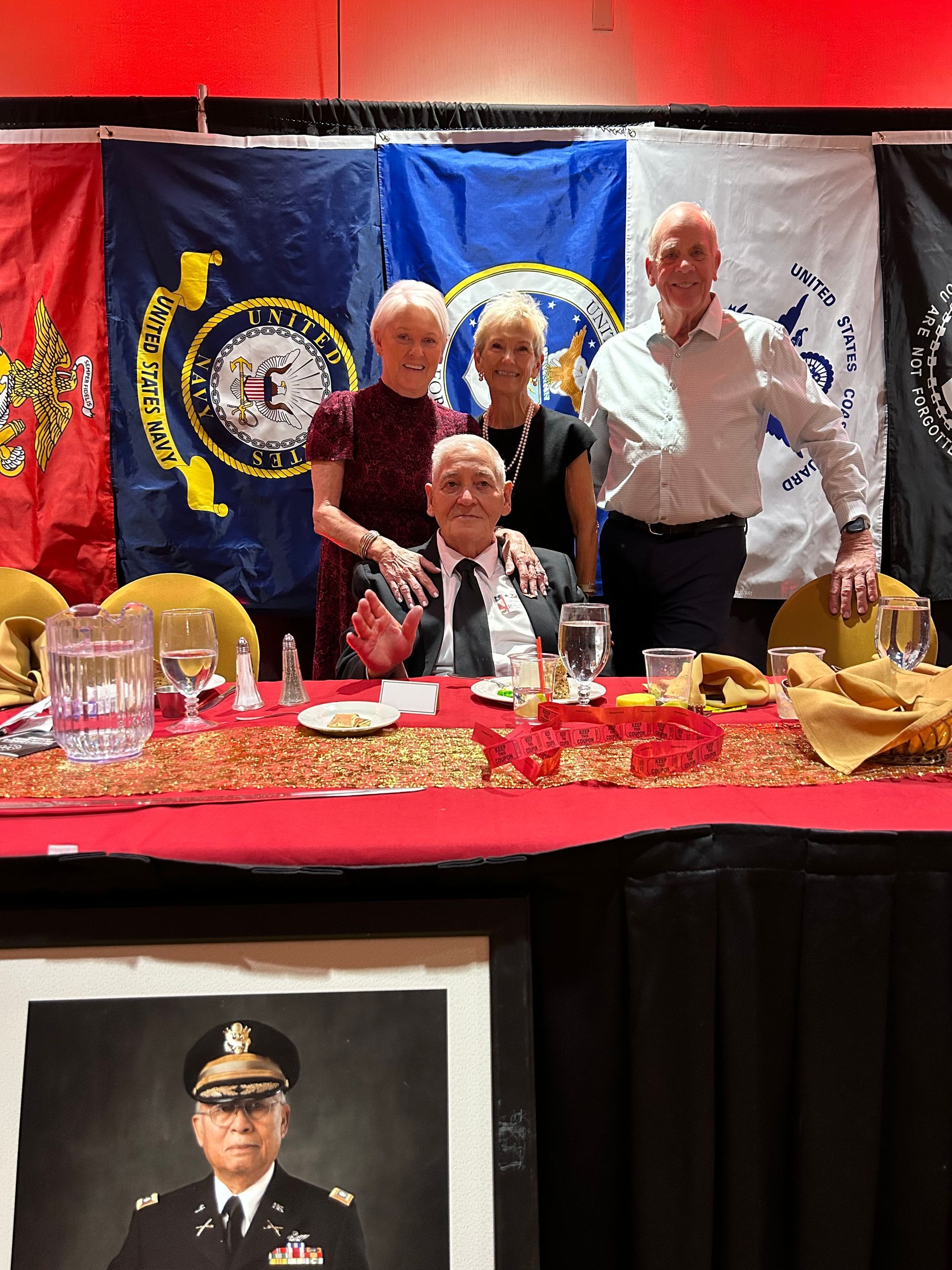 Three people pose behind a red banquet table with flags and a portrait in the background.