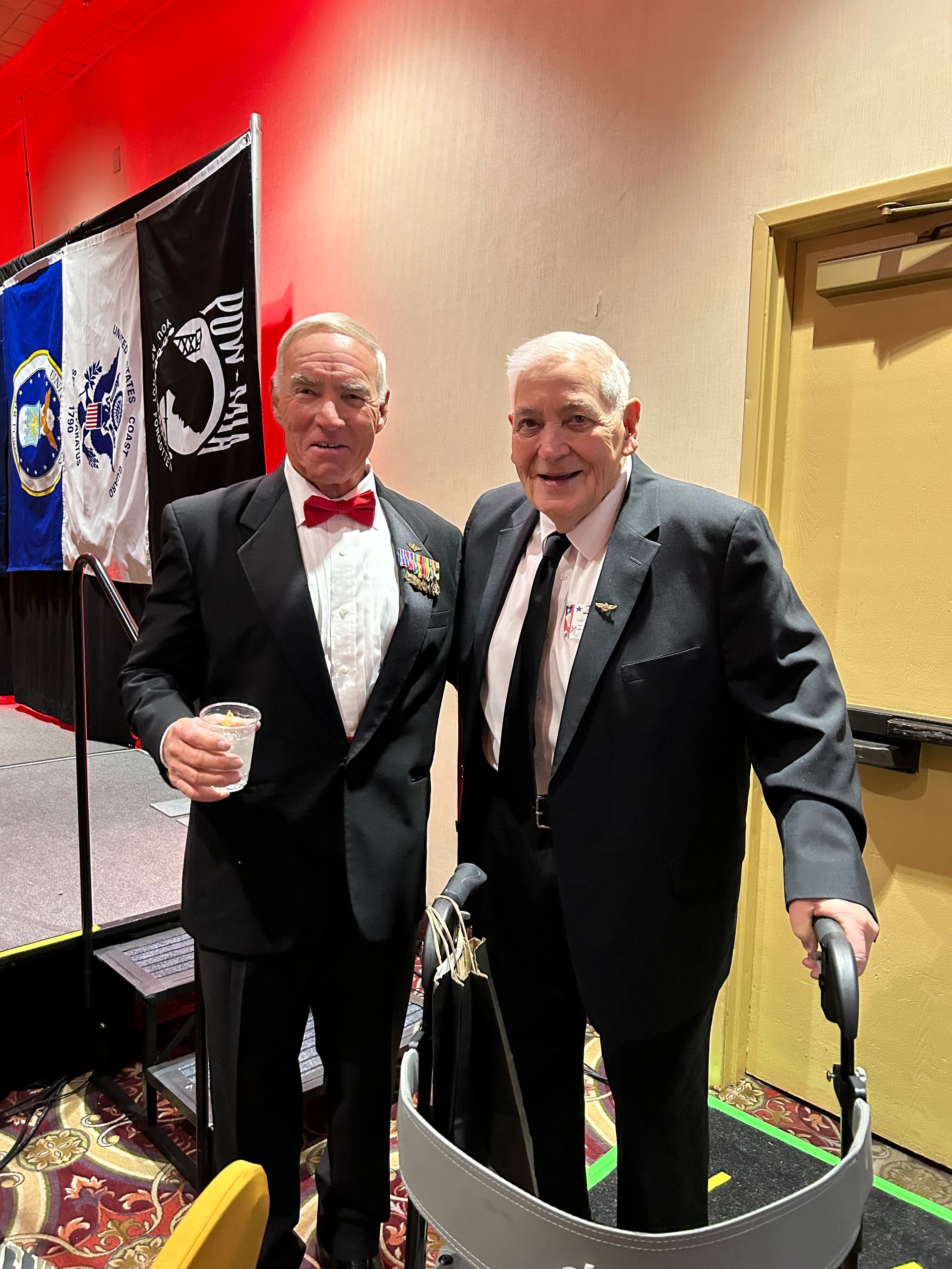 Two men in suits and bow ties pose together at an indoor event, with flags and a doorway behind them.